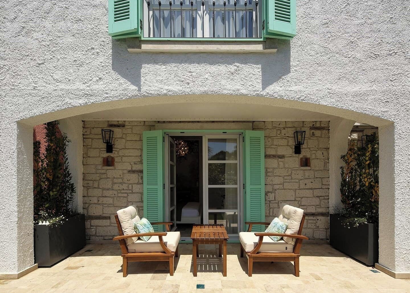 An outdoor seating area with two wooden chairs and a table, a stone wall, and a window with green shutters.