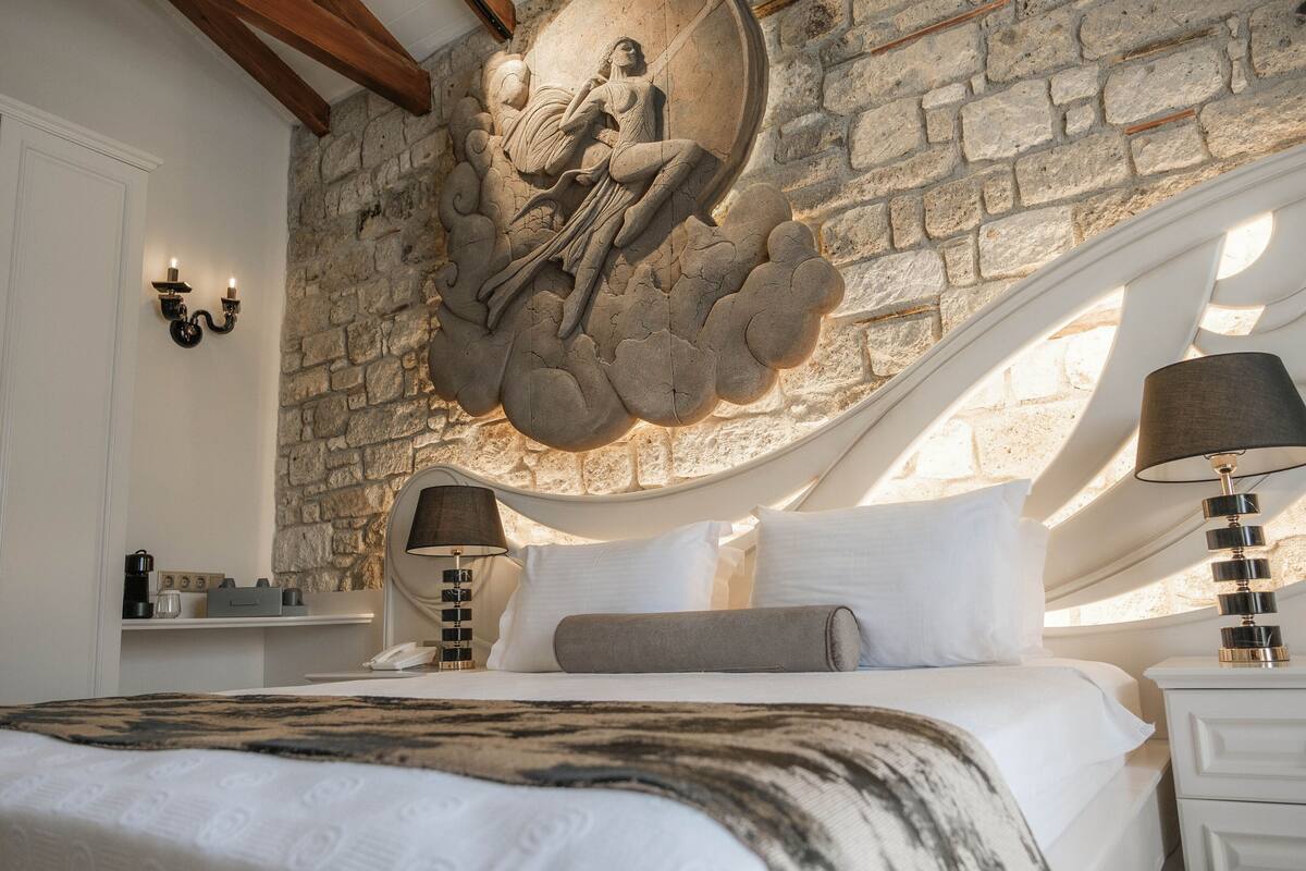 A bedroom with a stone wall, a bed with white bedding, a circular stone carving, and two bedside tables with lamps.