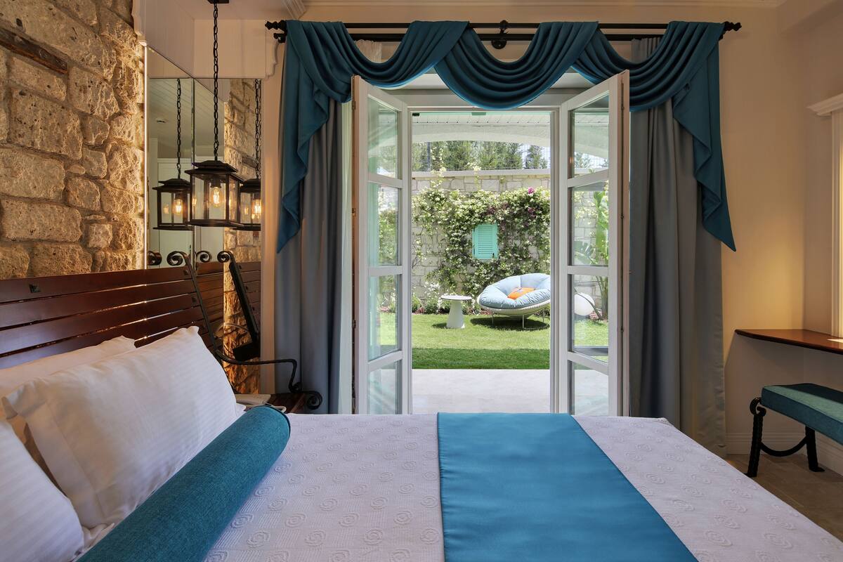 A bedroom with a large bed, a stone wall, a hanging light fixture, and a view of a garden through an open door.
