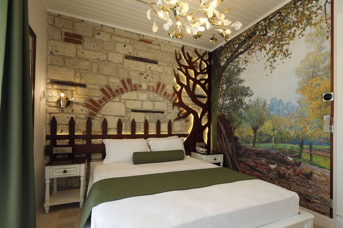 A bedroom with a brick wall, a wooden tree design, a bed with a green throw, and a mural of a landscape.