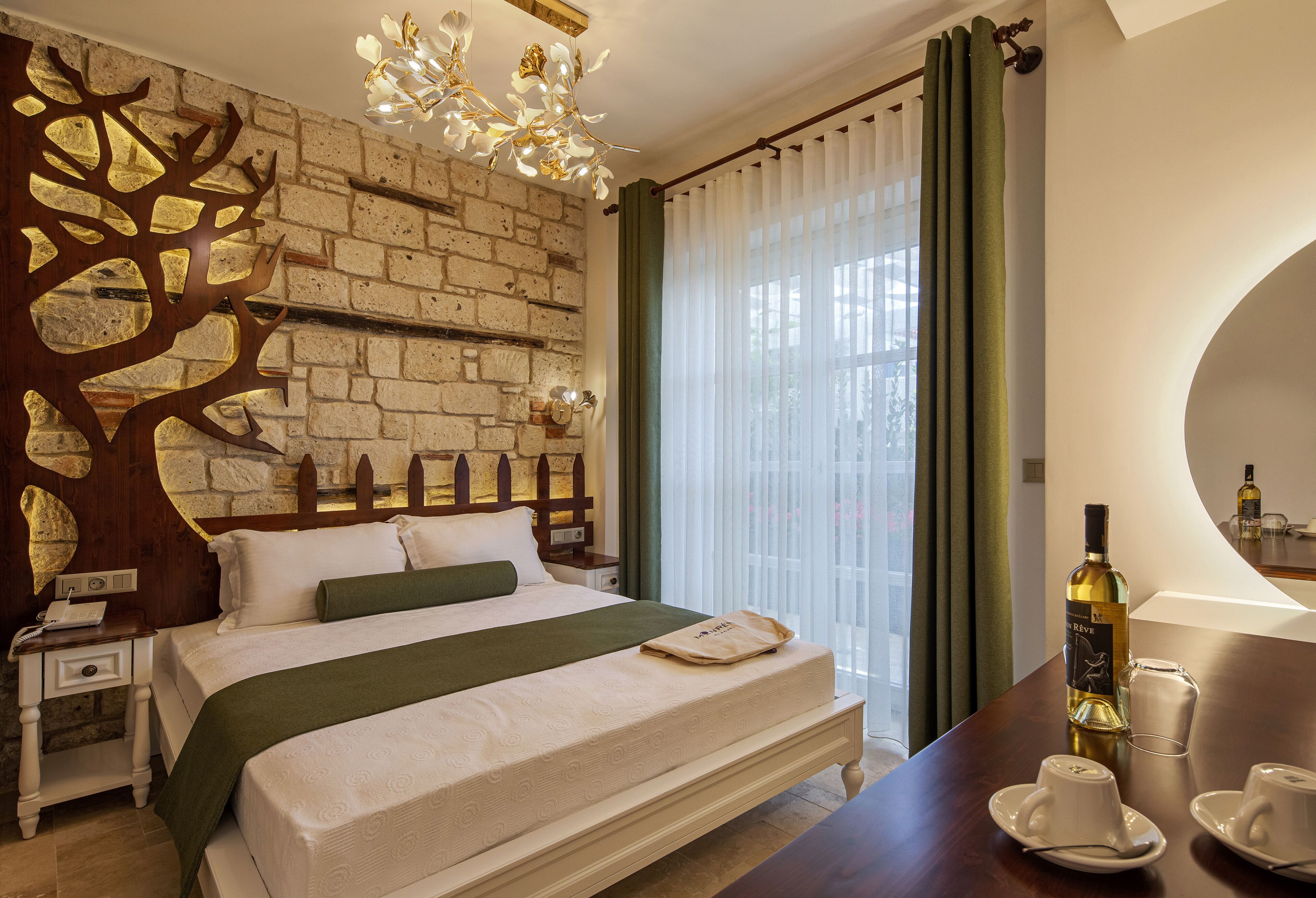 Habitación de hotel con pared de ladrillo, cama con manta verde, cabecero de madera, mesita de noche, lámpara, botella de vino y una copa sobre la mesa.