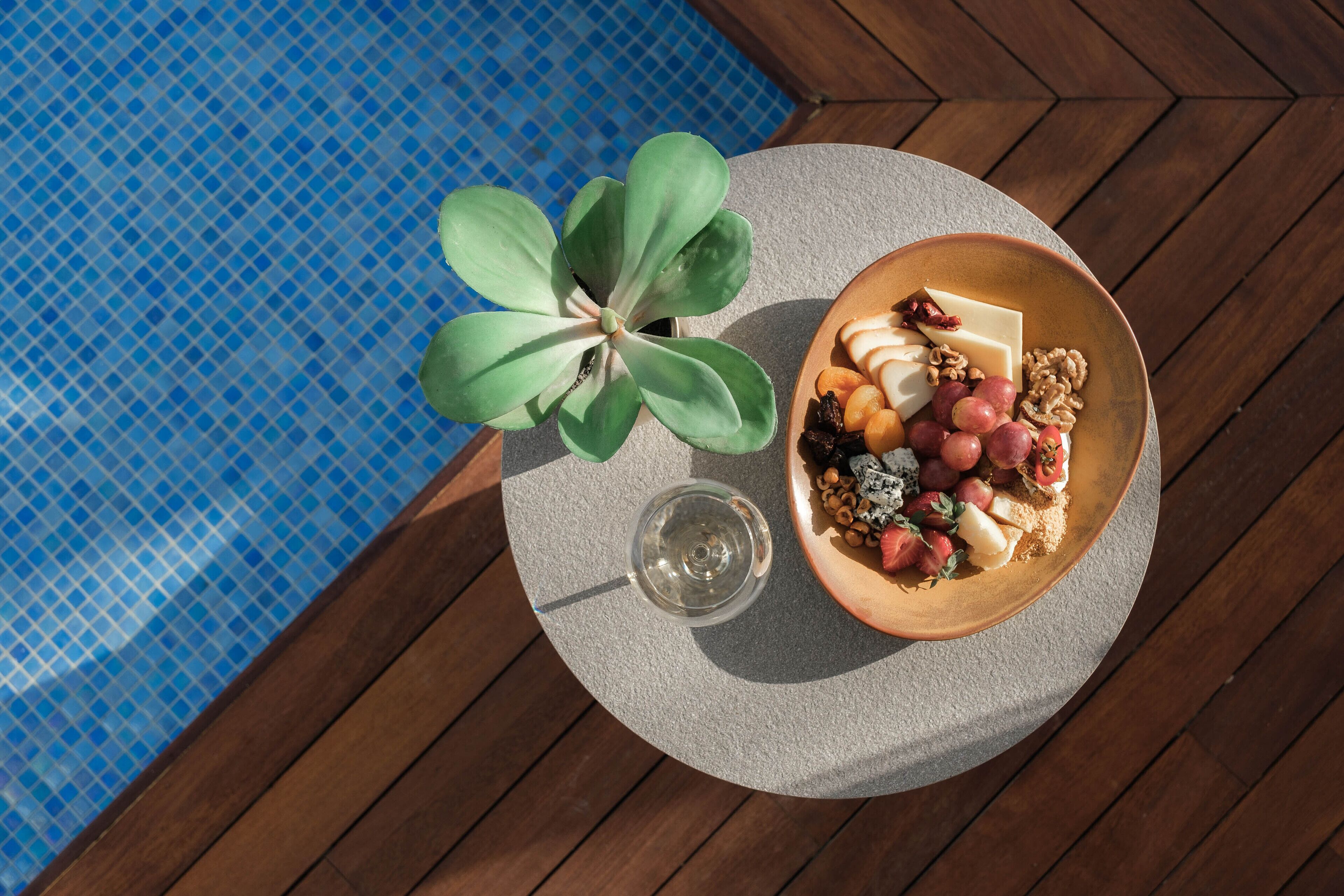 A round table with a bowl of food, a glass of wine, and a potted plant by a pool.