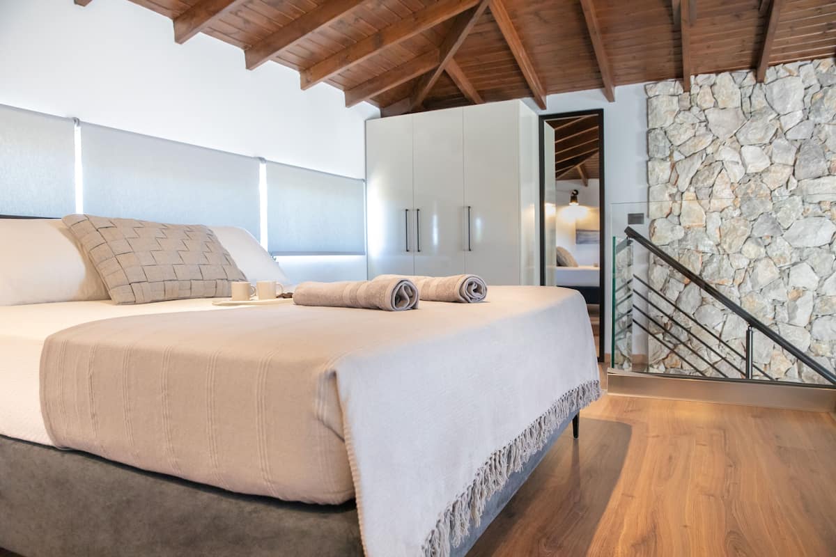 A bedroom with a large bed, wooden ceiling beams, a stone wall, and a staircase.