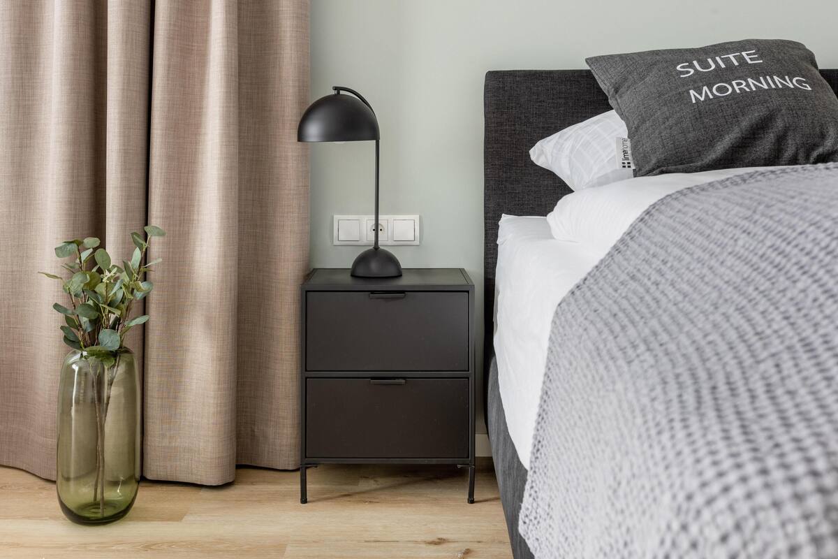 Ein ordentlich eingerichtetes Schlafzimmer mit Bett, Nachttisch, Lampe und einer Vase mit Grünpflanzen.
