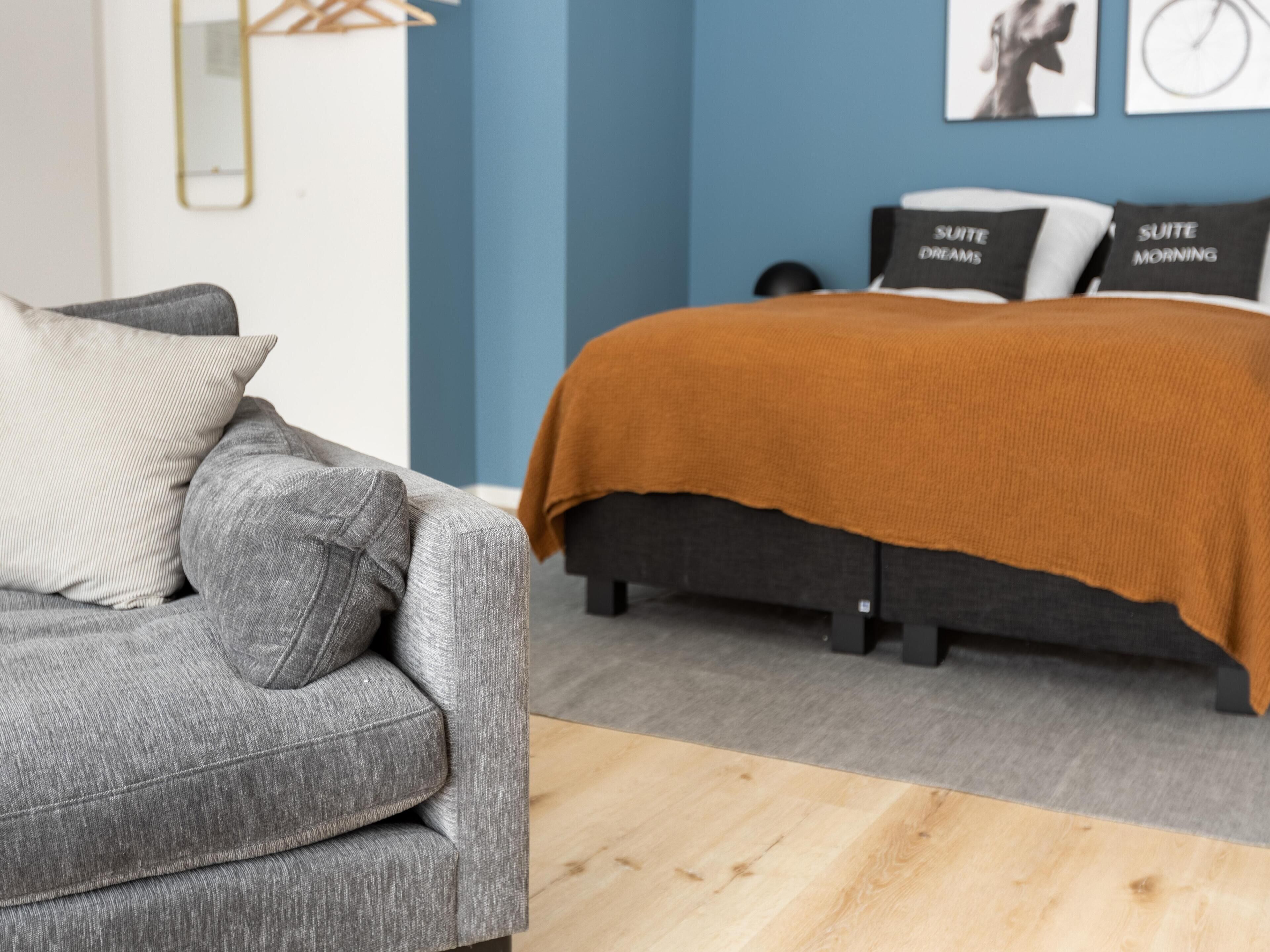 Ein modernes Schlafzimmer mit einem grauen Sofa, einem schwarzen Bett mit einer braunen Decke und zwei Kissen mit Text.