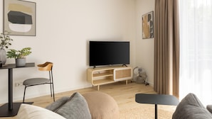 A modern living room with a sofa, a TV, a wooden entertainment unit, a small table, and a plant. - Limehome Osnabrück Johannisstraße (Osnabrueck)