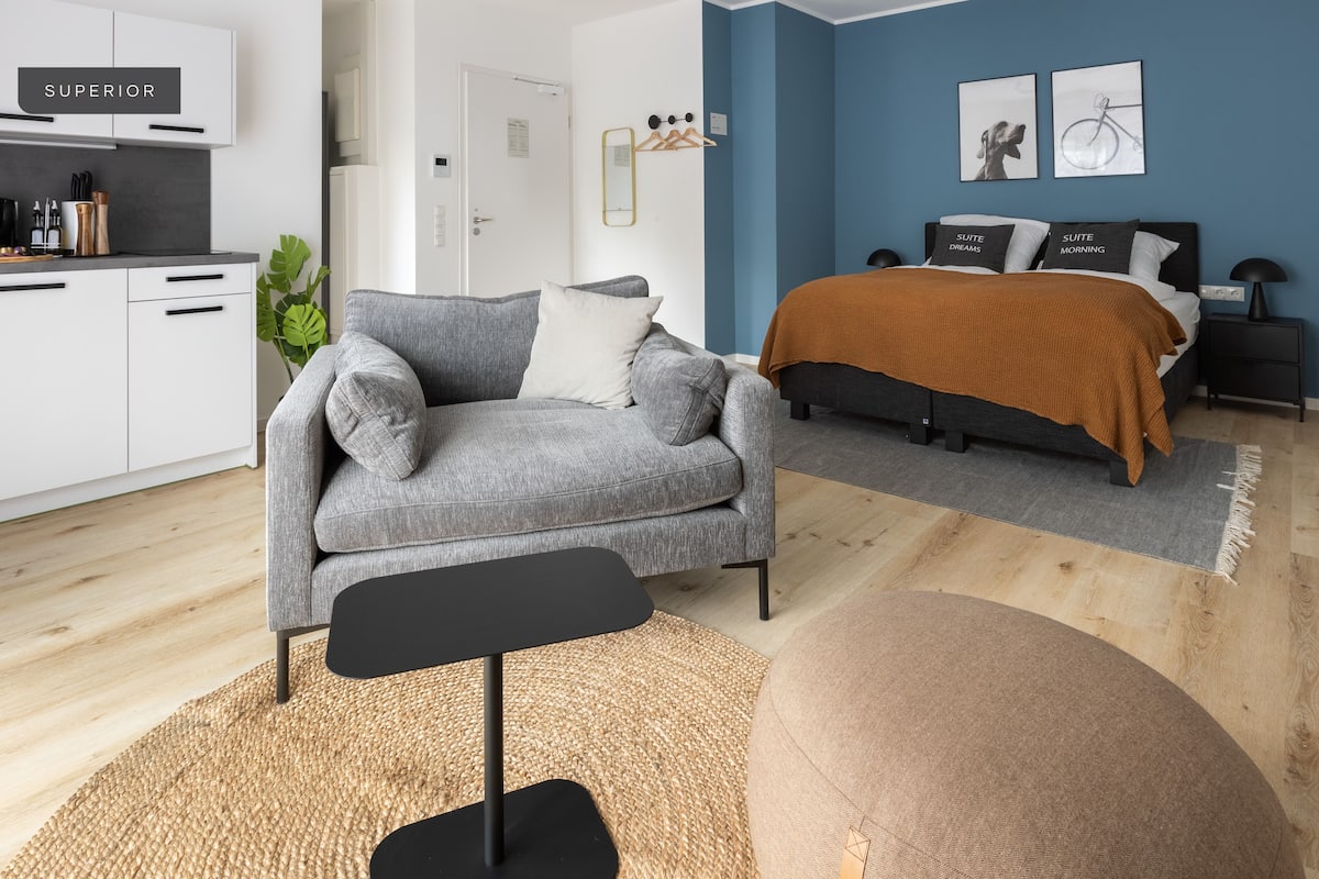 Ein modernes Hotelzimmer mit grauem Sofa, einem schwarzen Beistelltisch, einem braunen Hocker und einem Bett mit brauner Decke, sowie einer blauen Akzentwand.