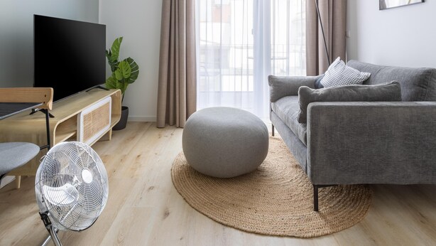 A modern living room with a grey sofa, a round ottoman, a flat-screen TV, a floor lamp, and a large window with curtains. - Limehome Osnabrück Johannisstraße (Osnabrueck)