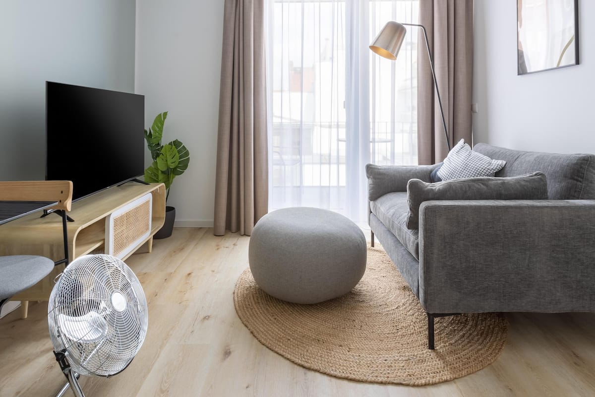 Ein modernes Wohnzimmer mit einem grauen Sofa, einem runden Hocker, einem Flachbildfernseher, einer Stehlampe und einem großen Fenster mit Vorhängen.