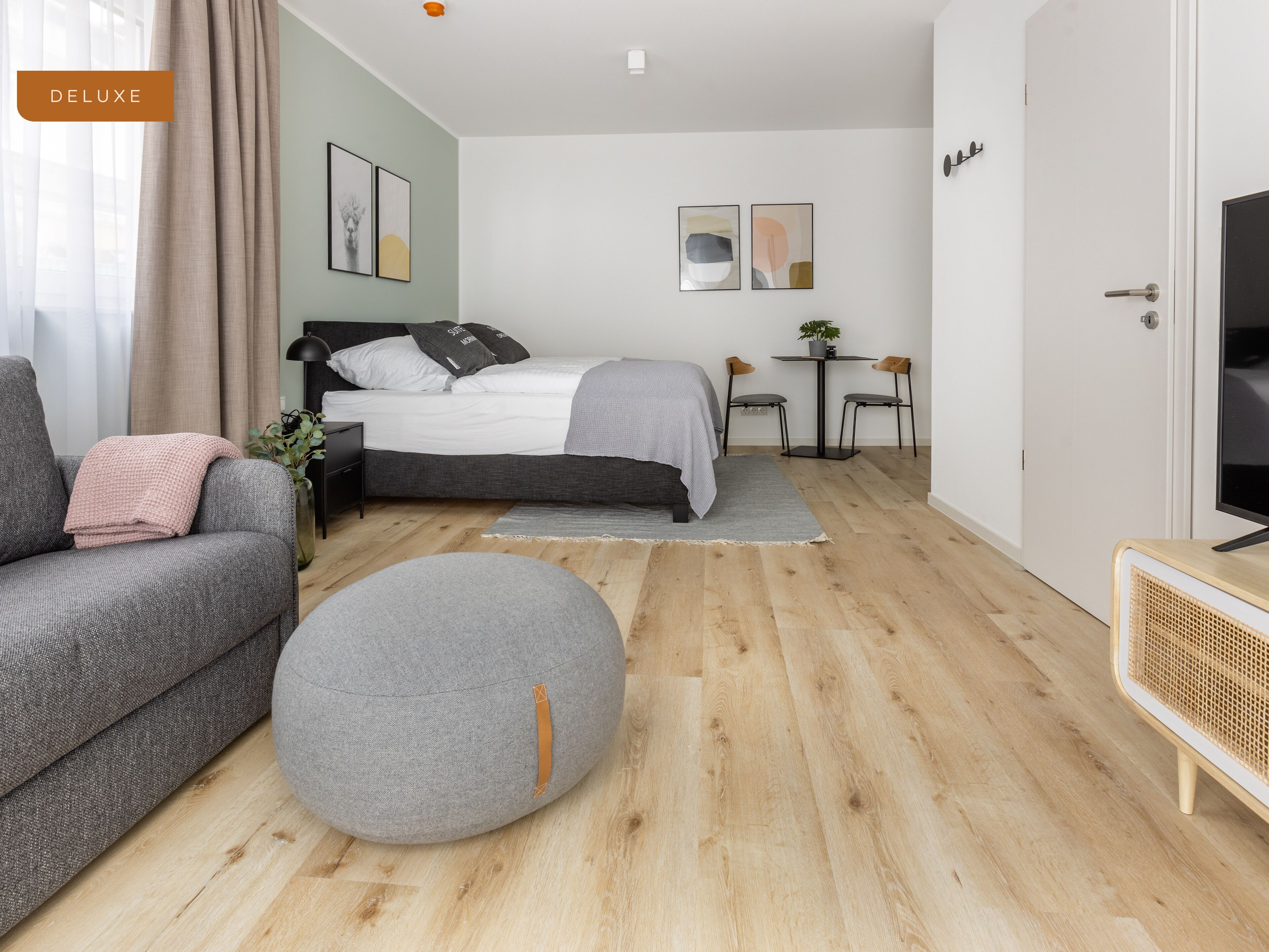 Un dormitorio moderno con cama, sofá gris, reposapiés y televisor.