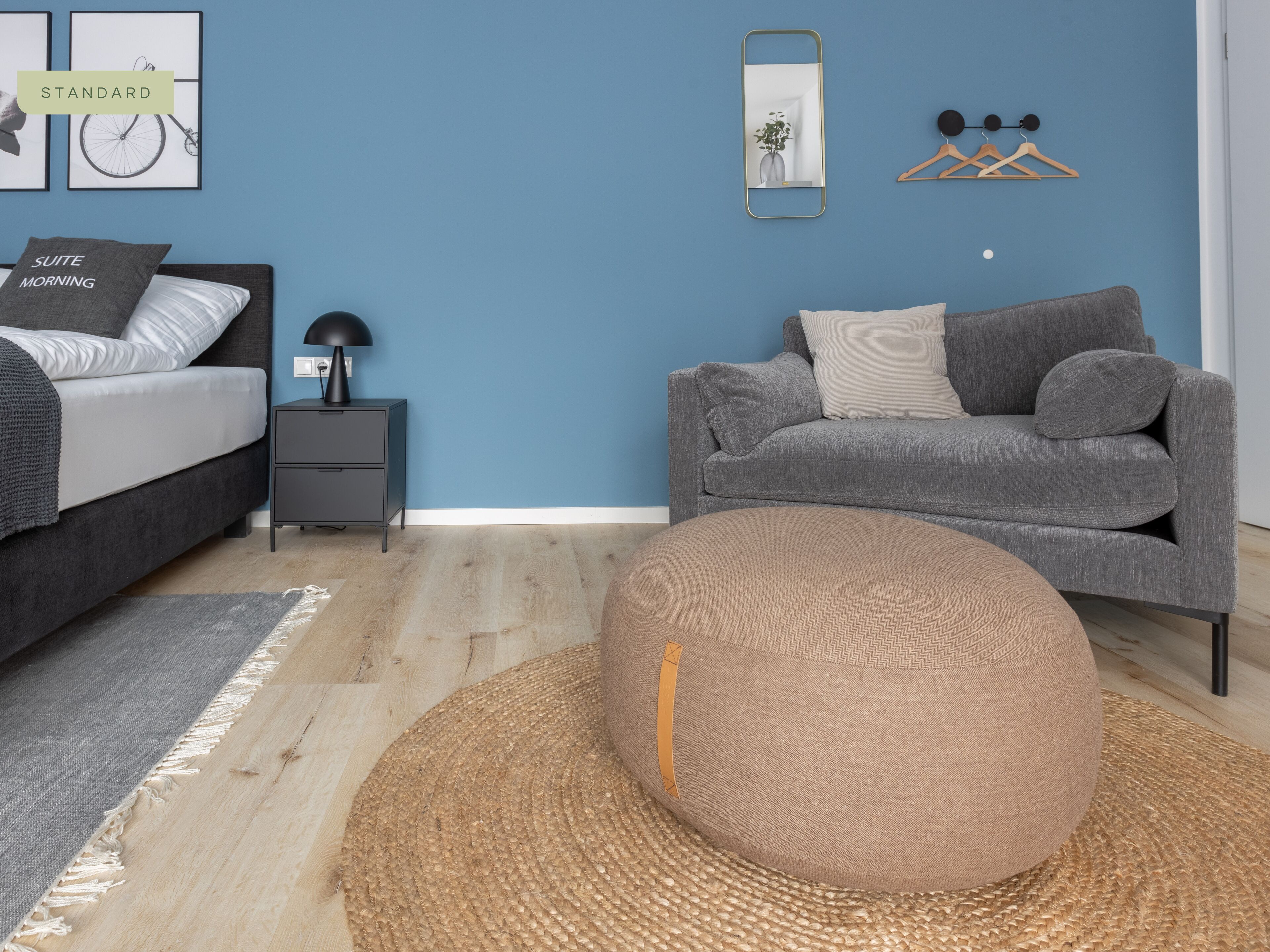 A modern bedroom with a bed, a grey sofa, a round ottoman, a nightstand, and a mirror on a blue wall.