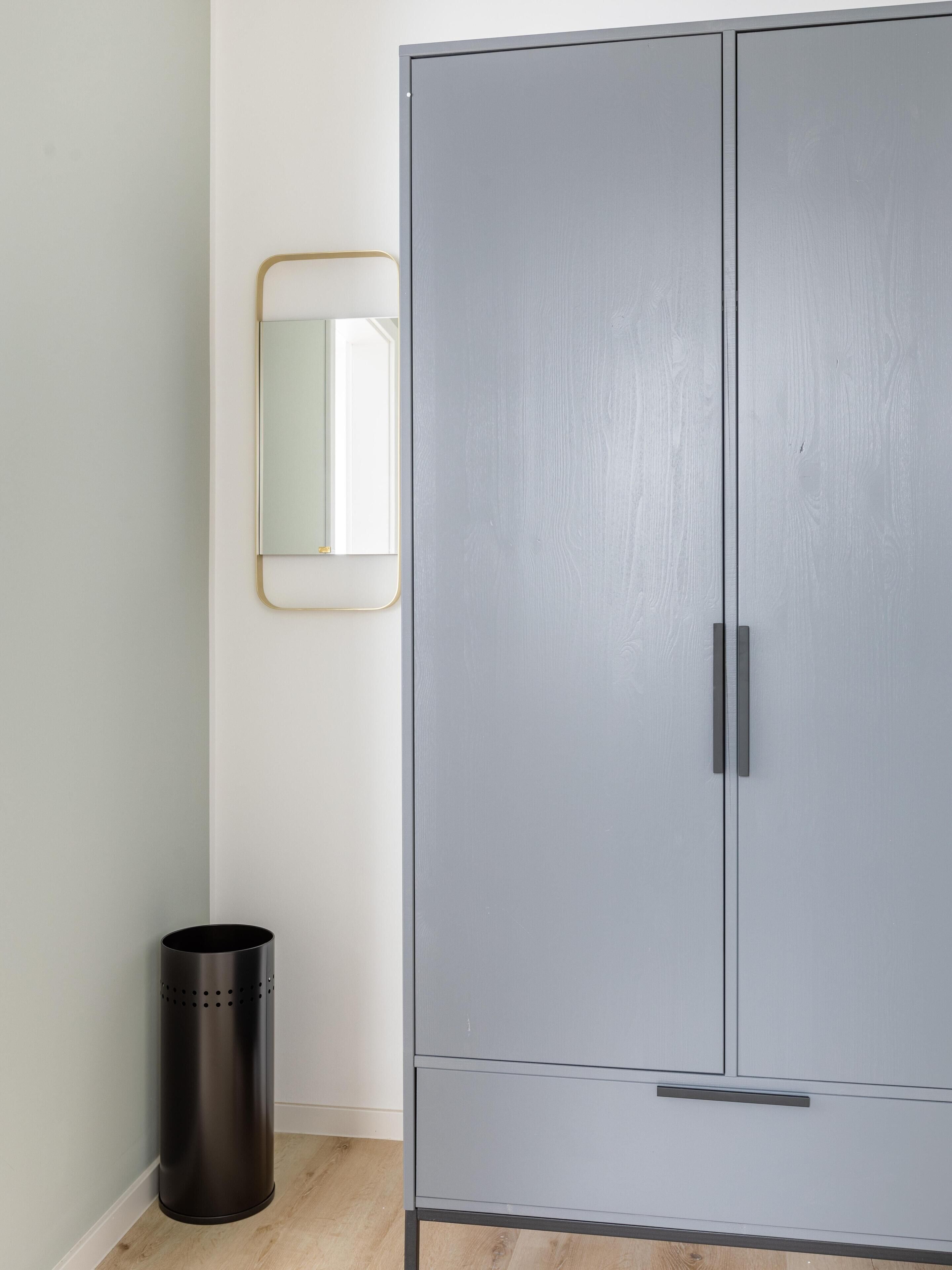 A grey wardrobe with two doors and a handle, a black trash bin, and a framed mirror on the wall.