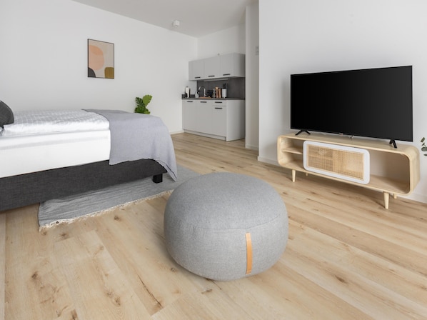 A modern bedroom with a bed, grey ottoman, TV, and kitchenette in the background.
