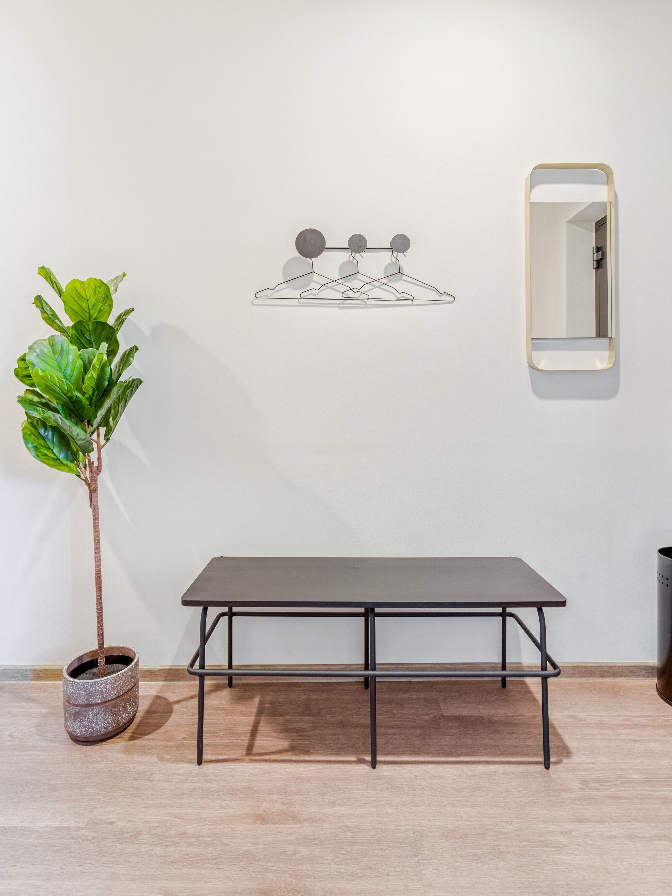 A minimalist room with a wooden floor, a table with metal legs, a potted plant, a wall-mounted mirror, and a simple art piece.