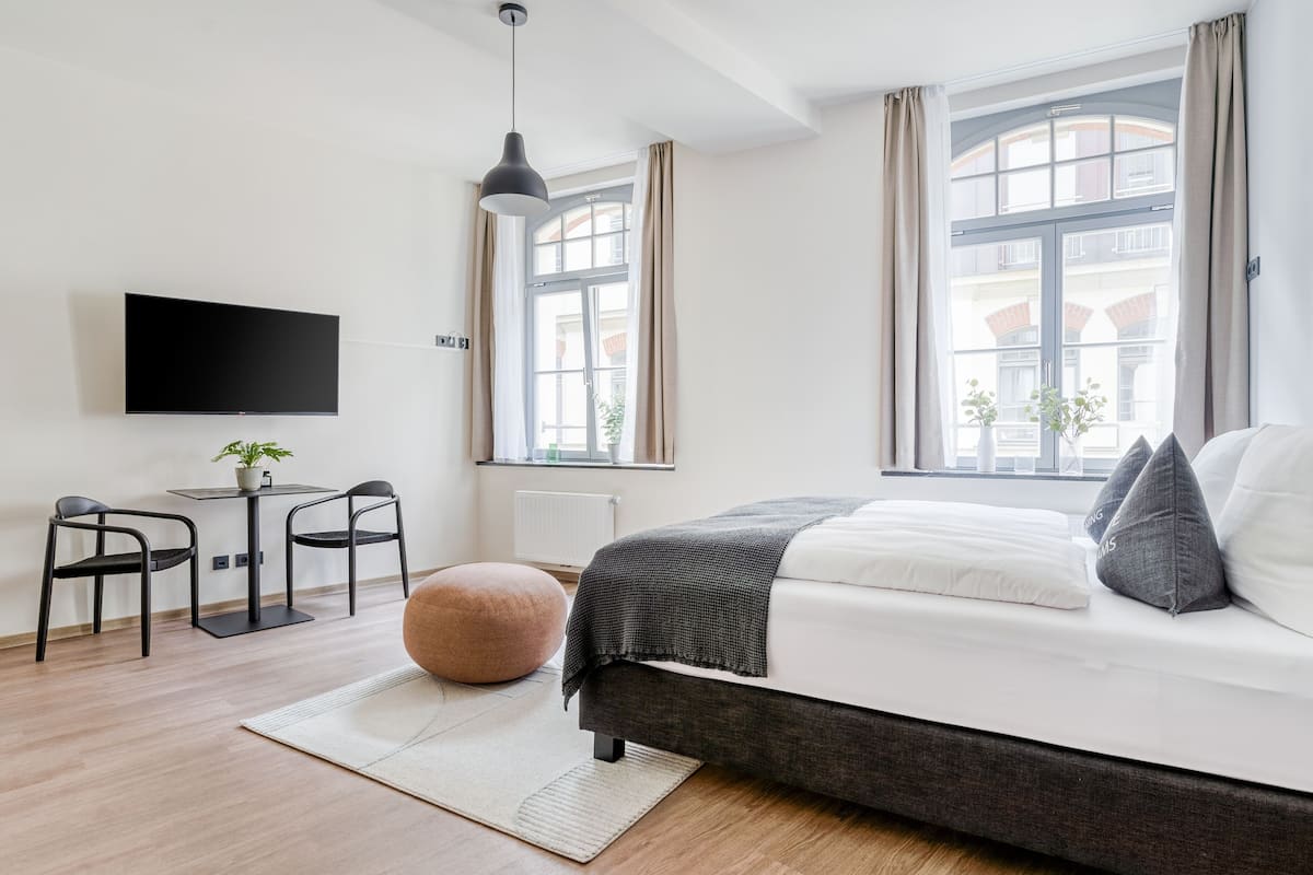 A modern bedroom with a bed, a desk, chairs, a television, and large windows.