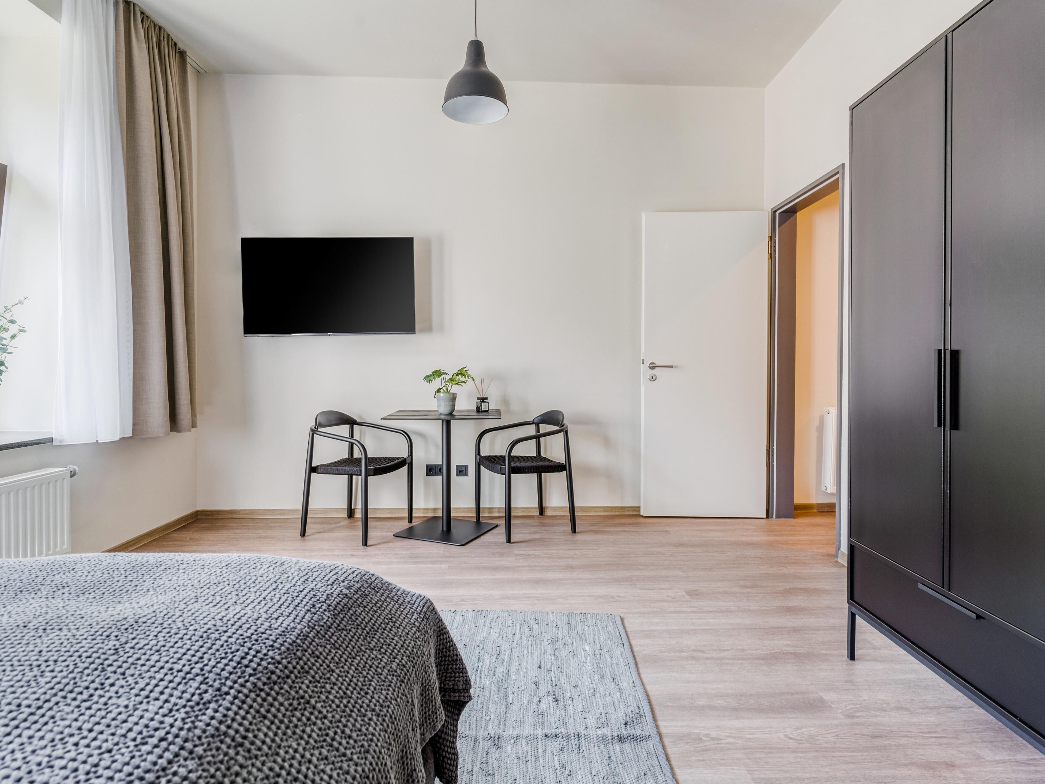 Un dormitorio moderno con cama, escritorio con dos sillas, televisor en la pared y armario.
