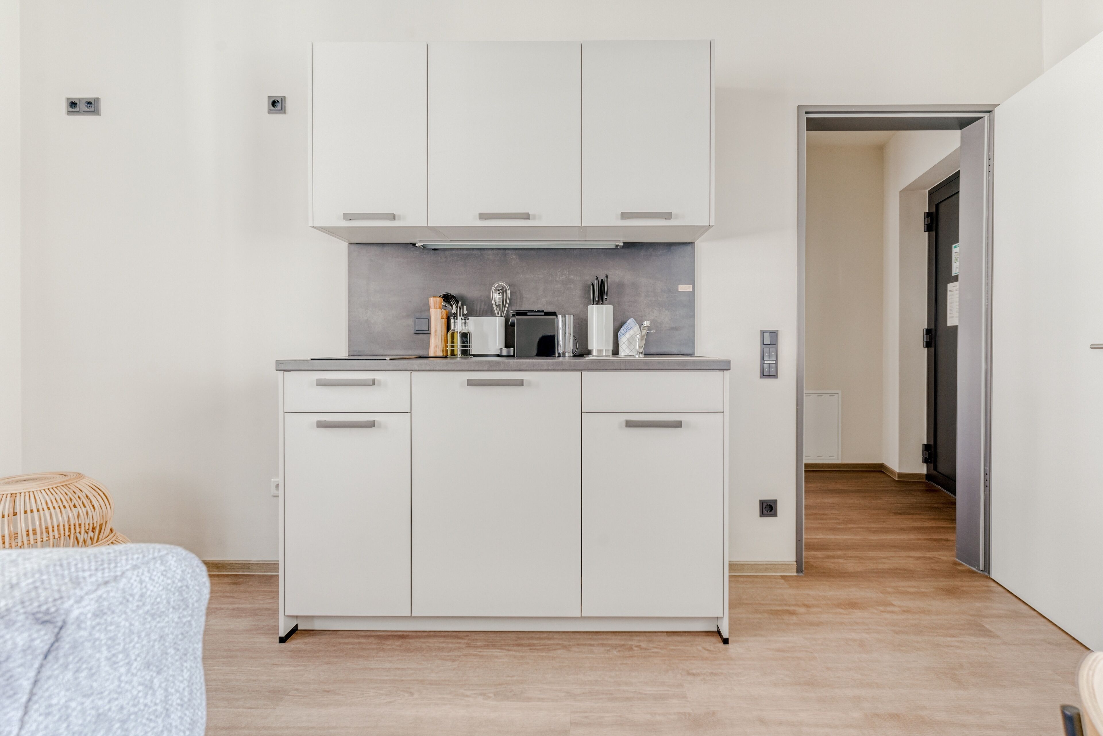 A compact kitchen with white cabinets, a countertop, and a toaster. A wicker chair is visible in the foreground.