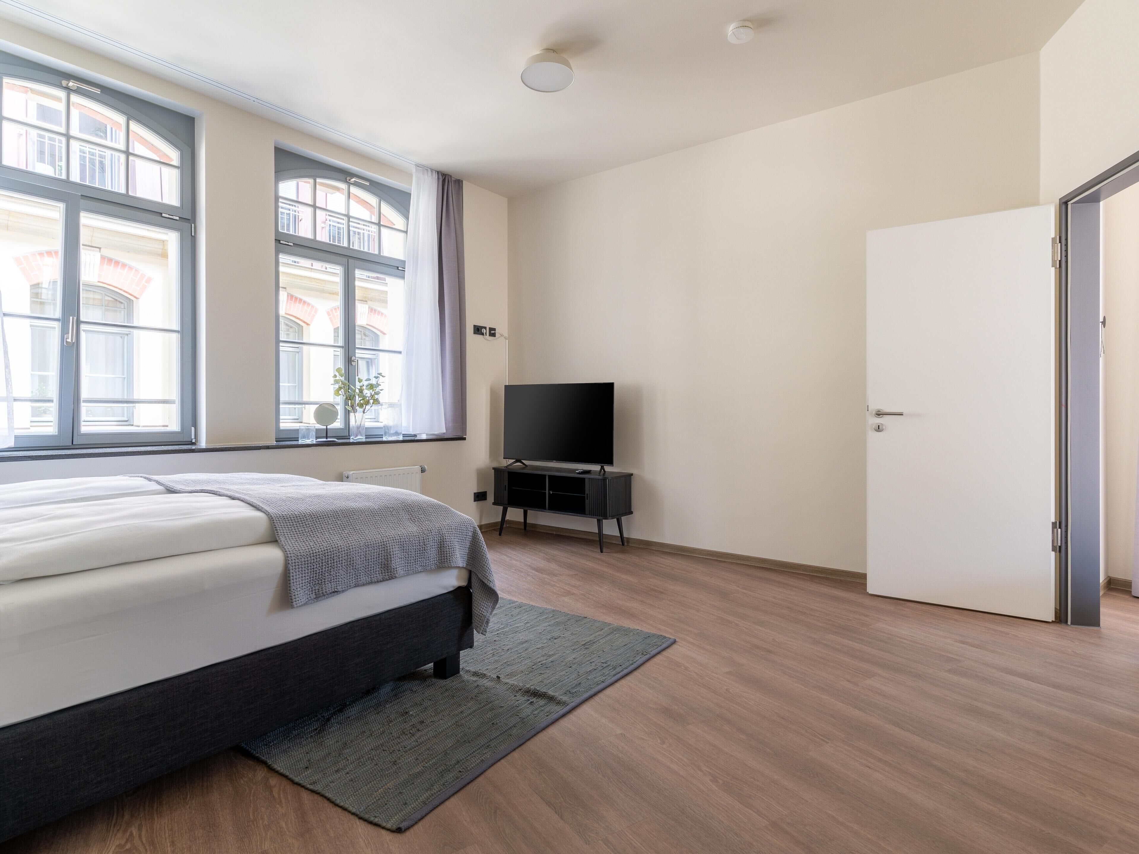 Un dormitorio moderno con cama, televisor sobre un mueble, ventana con cortinas y una puerta.