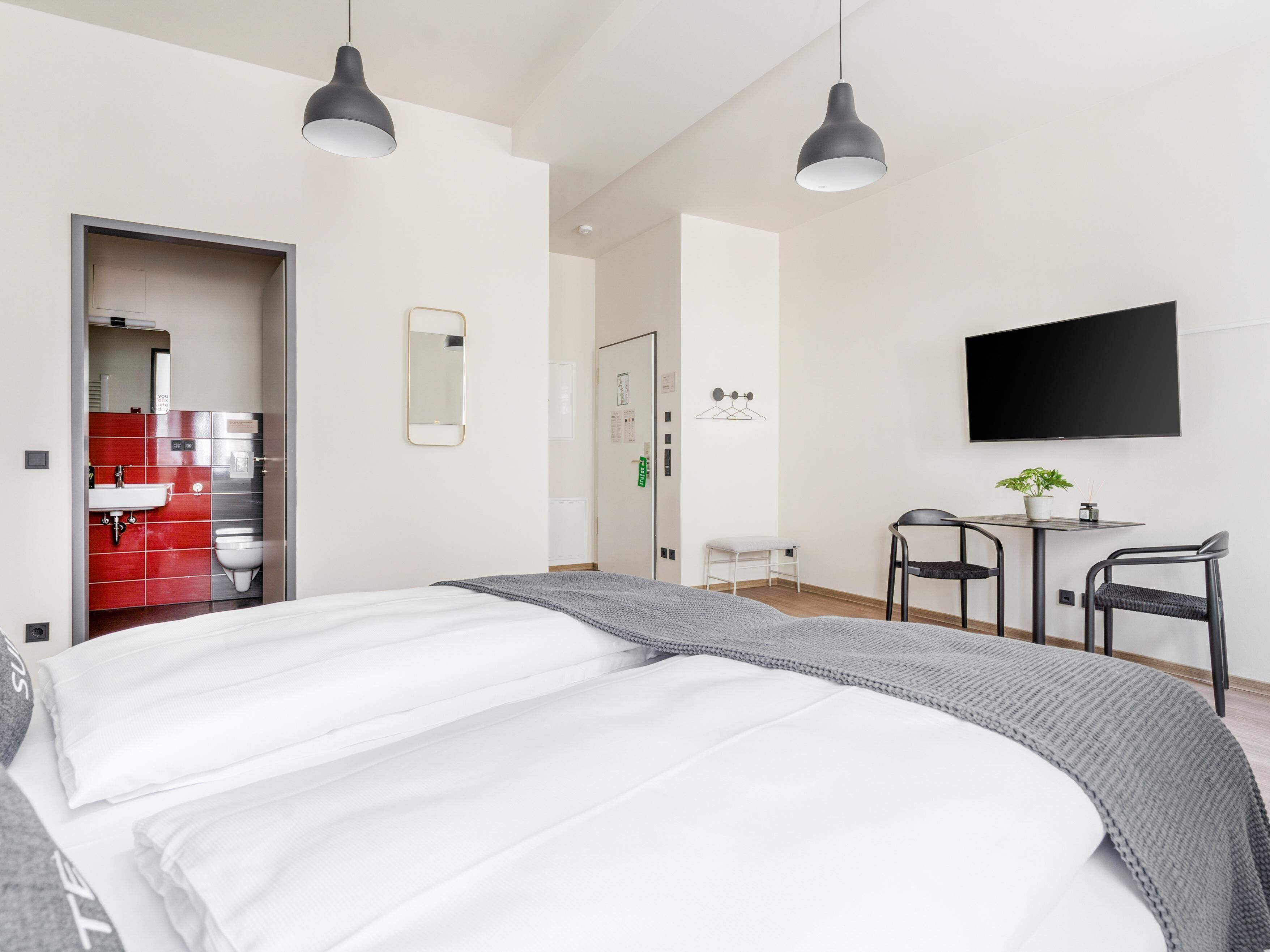Une chambre d’hôtel moderne, équipée d’un lit, de deux lampes suspendues, d’une télévision, d’une petite table avec des chaises et d’une salle de bain visible à travers une porte ouverte.