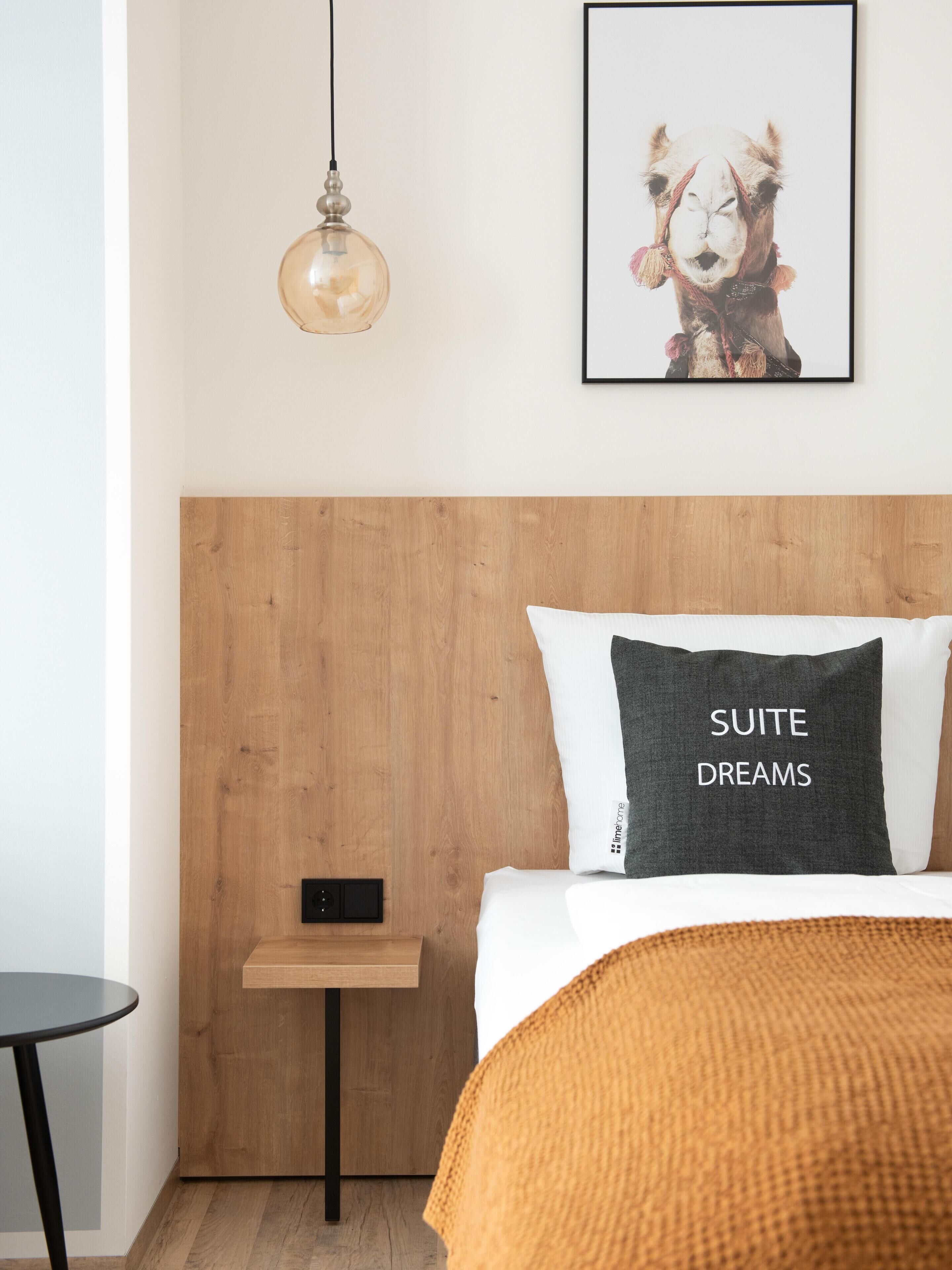 Ein modernes Schlafzimmer mit einem hölzernen Kopfteil, einem Nachttisch, einem gerahmten Bild an der Wand und einem Kissen mit dem Aufdruck 'SUITE DREAMS'.