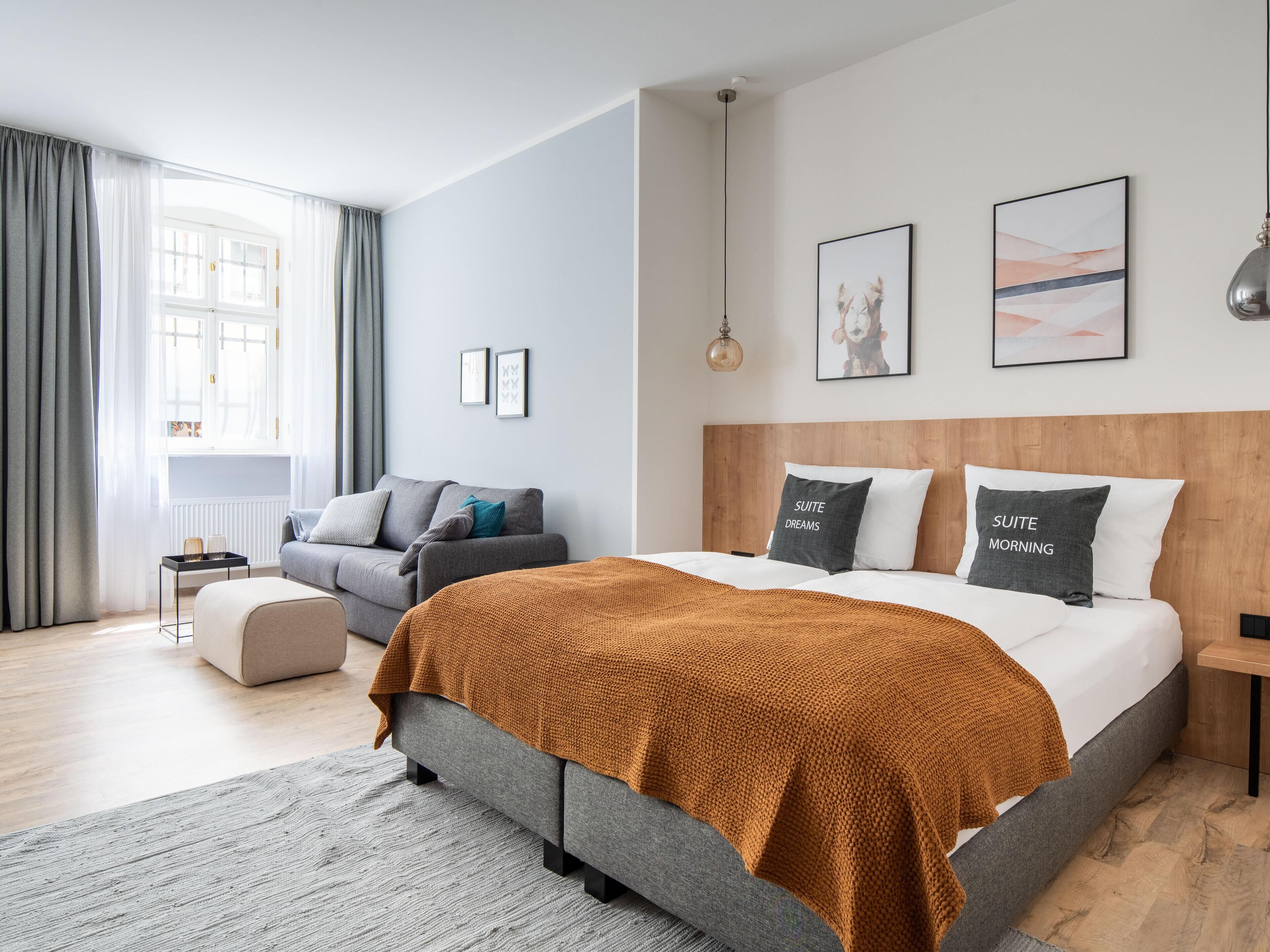 A modern bedroom with a large bed, a grey sofa, and a wooden headboard. There are two framed pictures on the wall and a window with curtains.