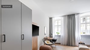 Una sala de estar moderna con una televisión de pantalla plana, piso de madera, un ventanal con cortinas, una silla de mimbre, un otoman redondo y un sofá gris.