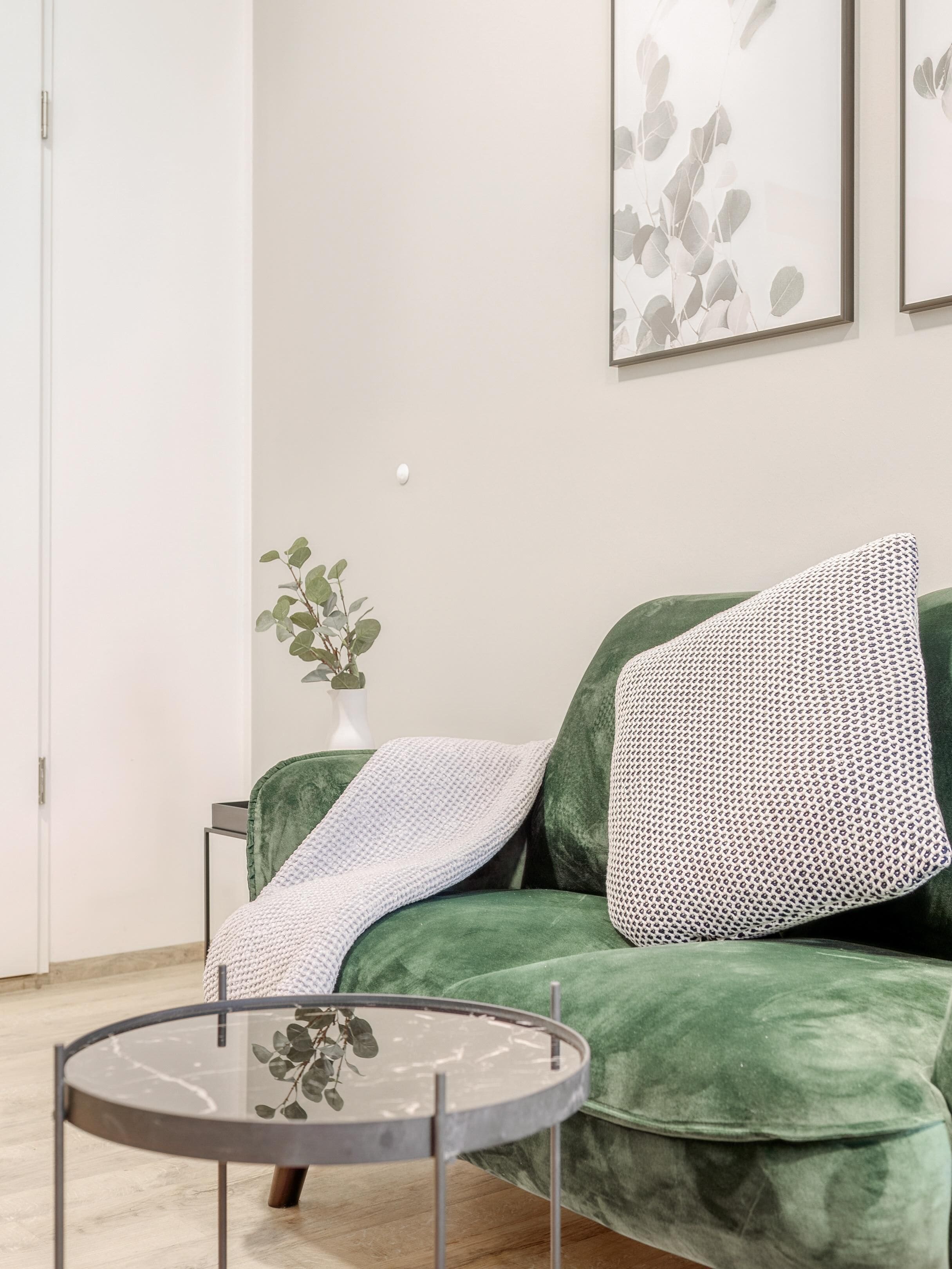 A green sofa with patterned pillows, a glass coffee table, a small plant, and framed artwork on the wall.