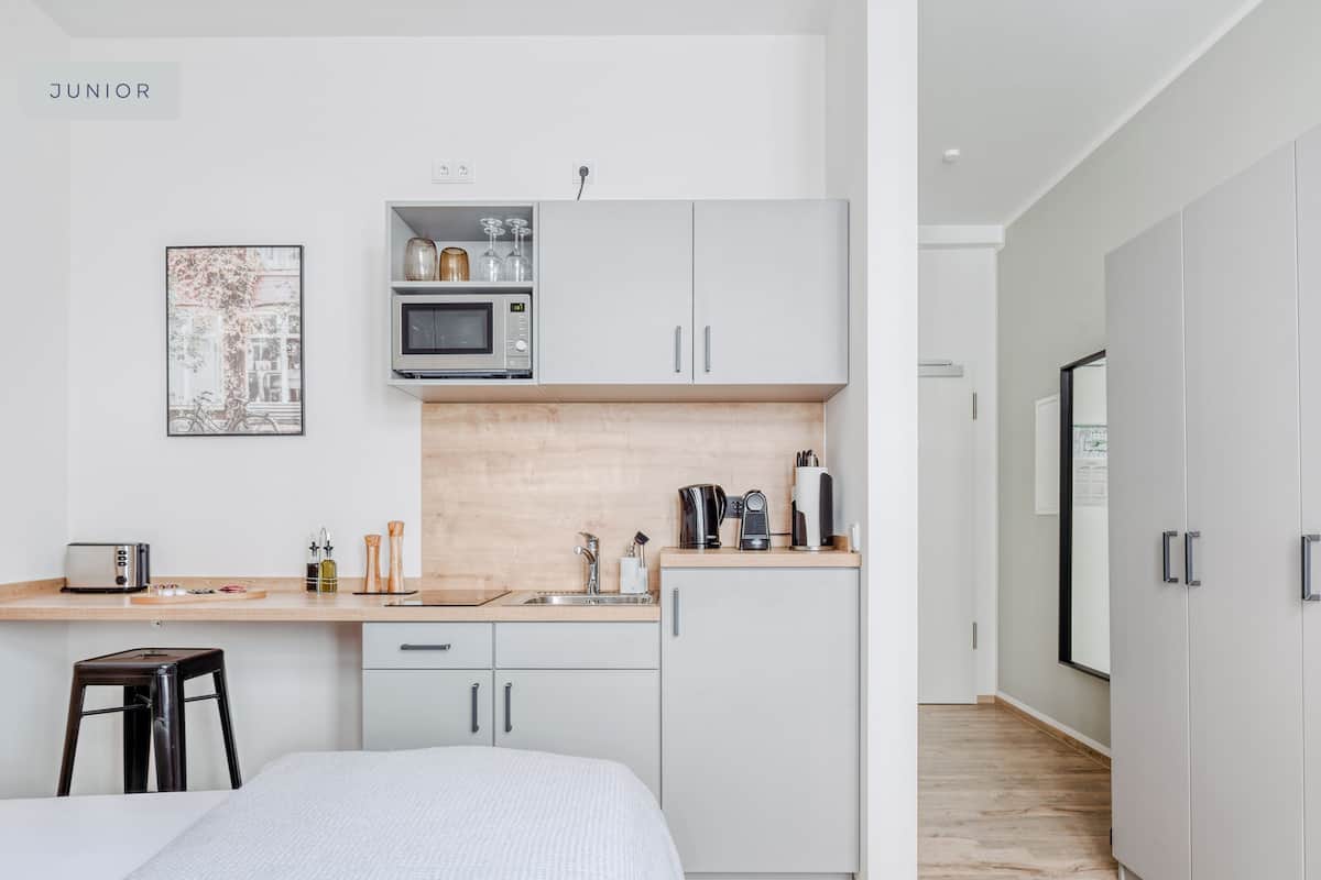 Een moderne, compacte slaapkamer met een bed, een klein bureau en een kitchenette met een magnetron en koffiezetapparaat.