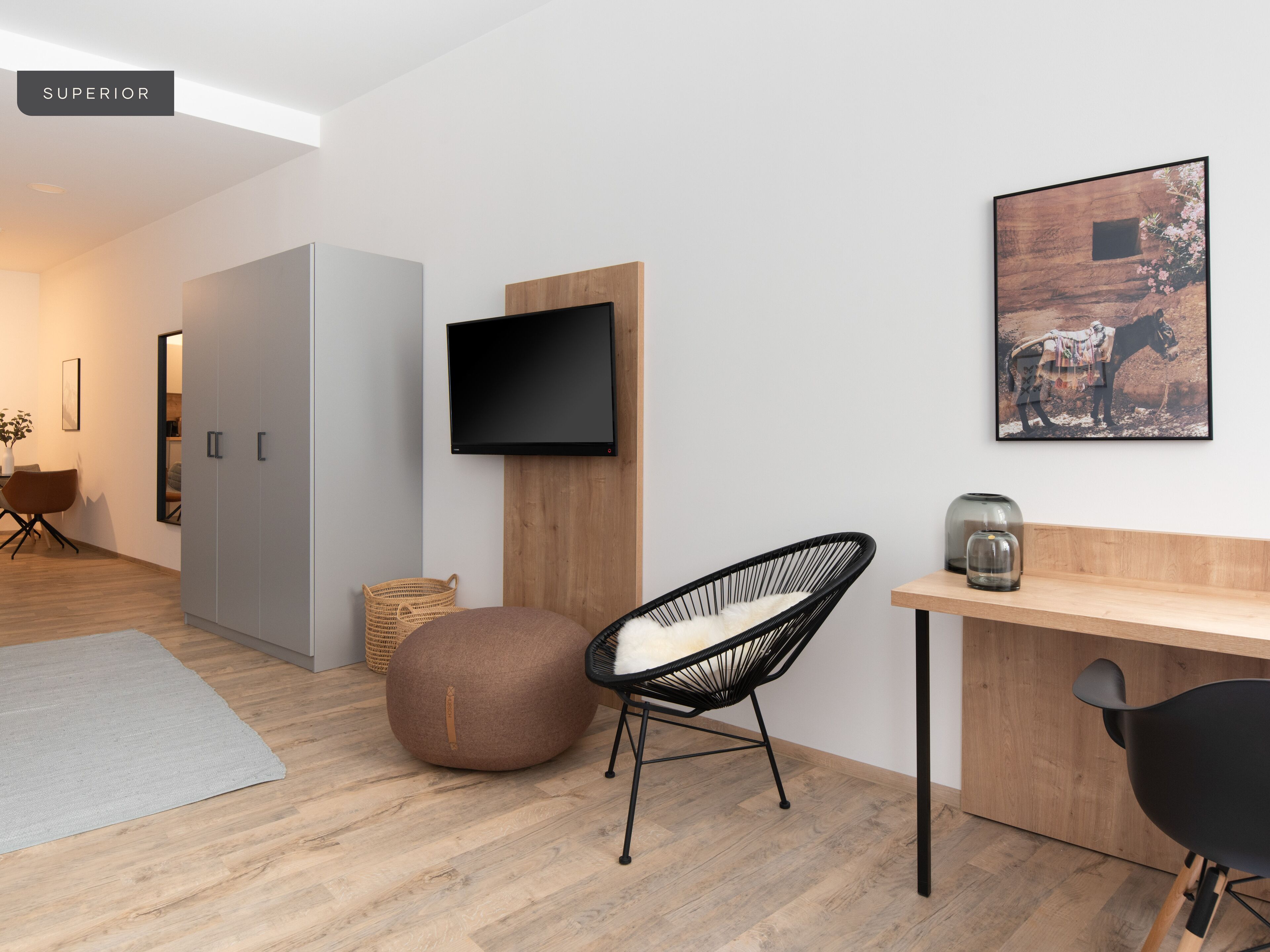 A modern living room with a flat-screen TV mounted on a wooden panel, a round bean bag chair, a black chair, a small table with a glass vase, and a framed picture on the wall.