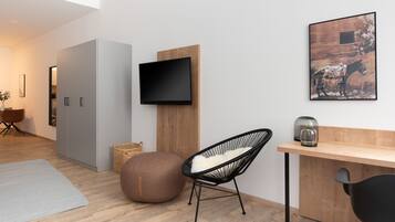 A modern living room with a flat-screen TV mounted on a wooden panel, a round bean bag chair, a black chair, a small table with a glass vase, and a framed picture on the wall.