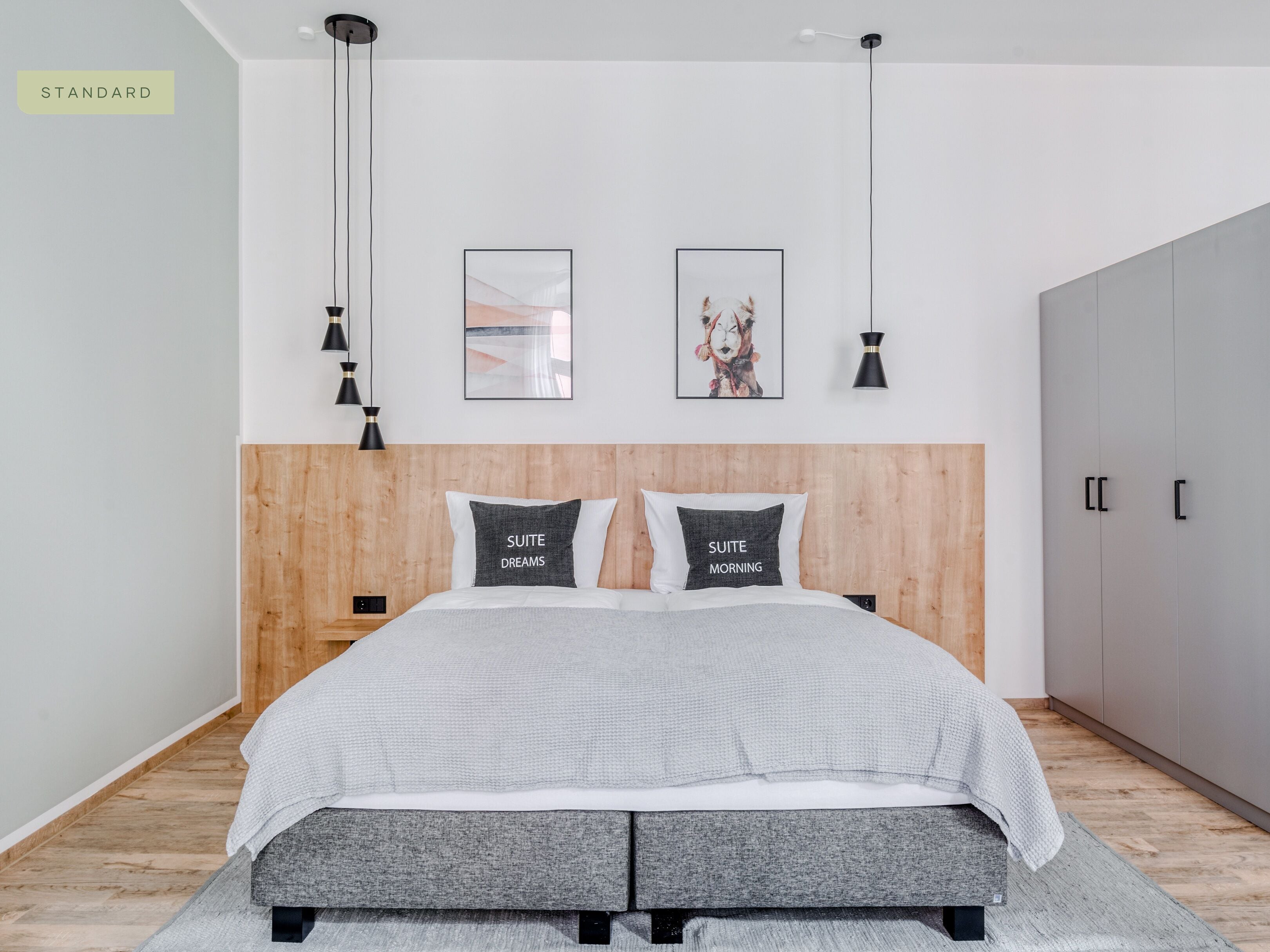 Une chambre moderne avec une tête de lit en bois, deux tables de chevet, une couvre-lit grise et deux lampes noires fixées au mur.