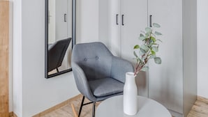 Una habitación moderna con un sillón gris, un jarrón blanco con una planta, una mesa redonda blanca y un armario gris.