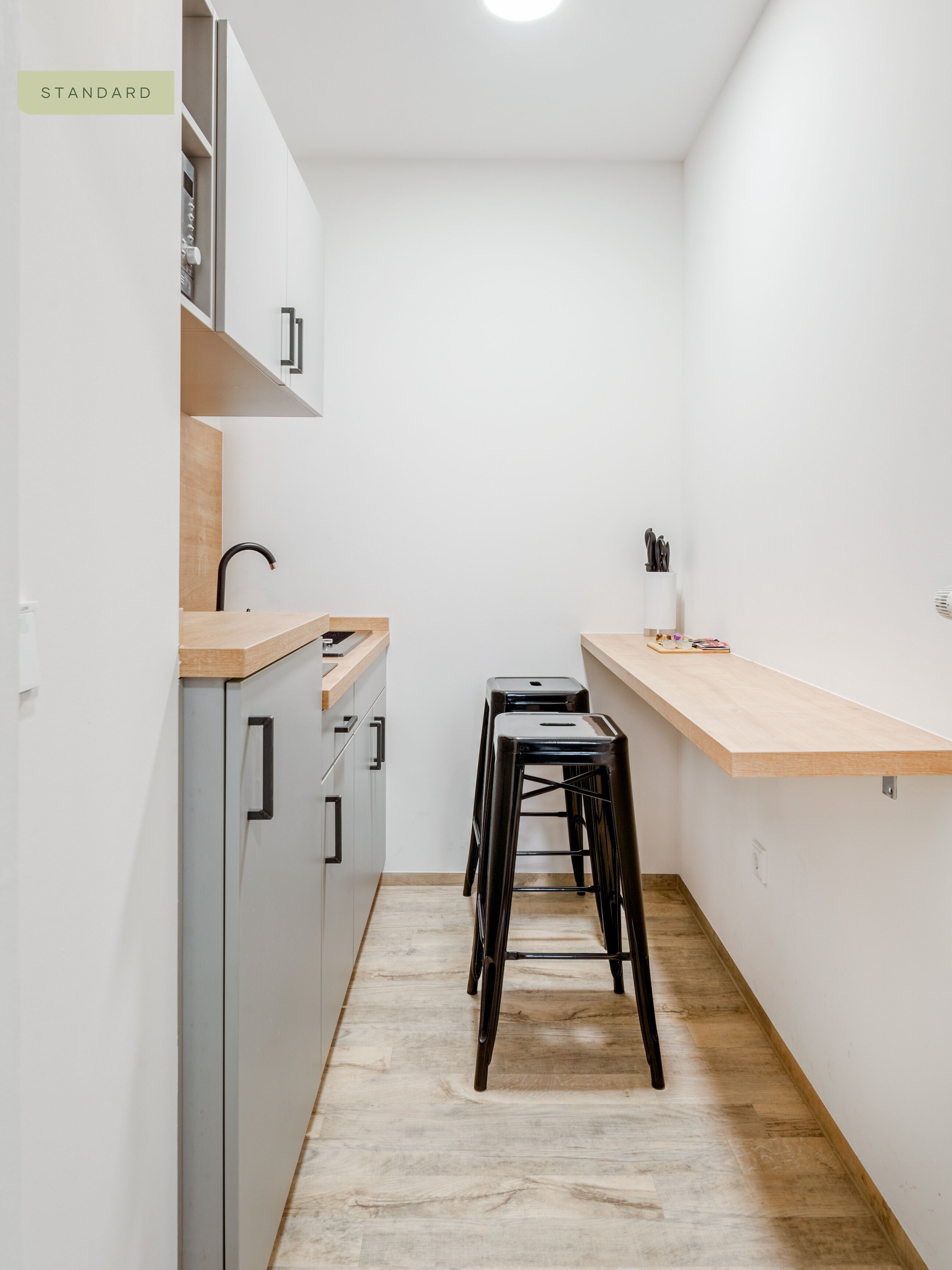 A narrow kitchen with a wooden countertop, two black stools, and a light-colored floor.