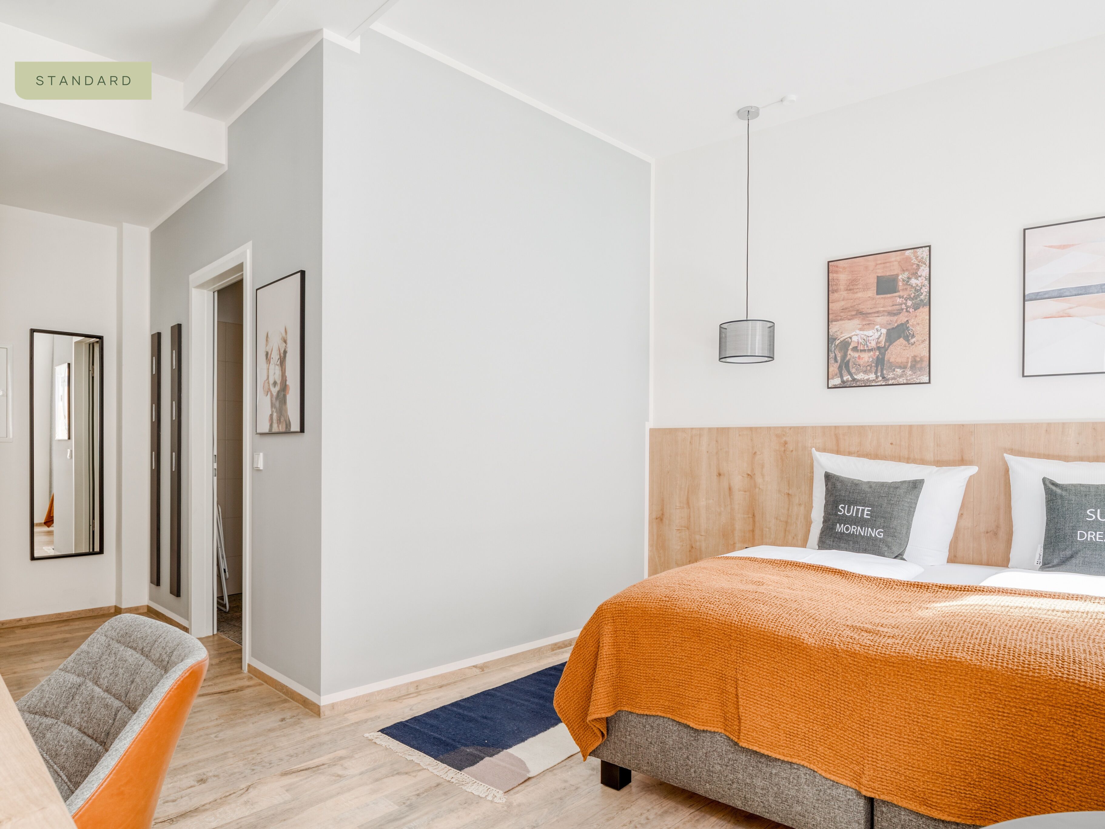 A modern bedroom with a bed, a wooden headboard, and a blue rug. There are framed pictures on the wall and a hanging light fixture.