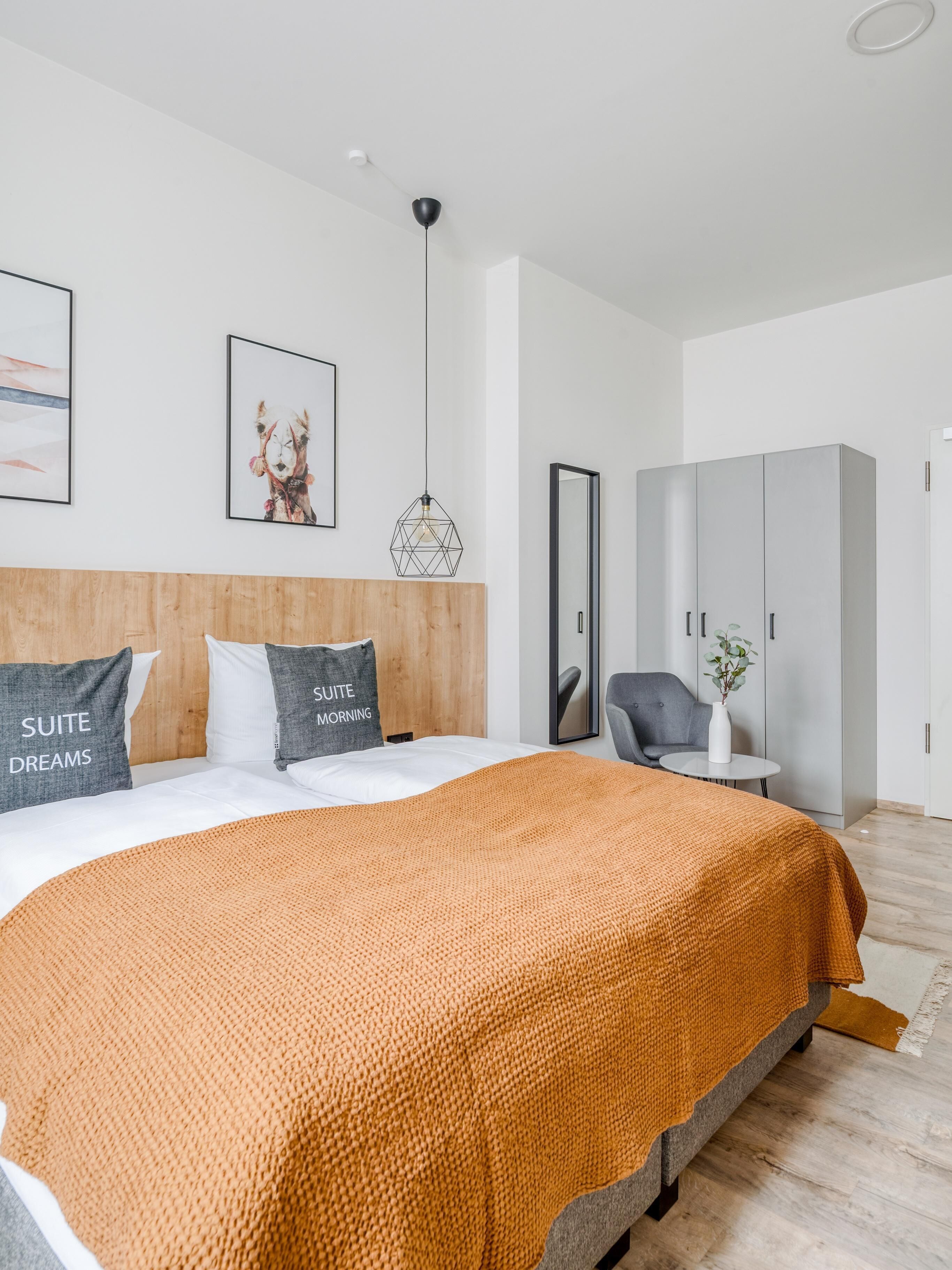 A modern hotel room with a large bed, a wooden headboard, a grey sofa, a small table with a plant, and a wardrobe.