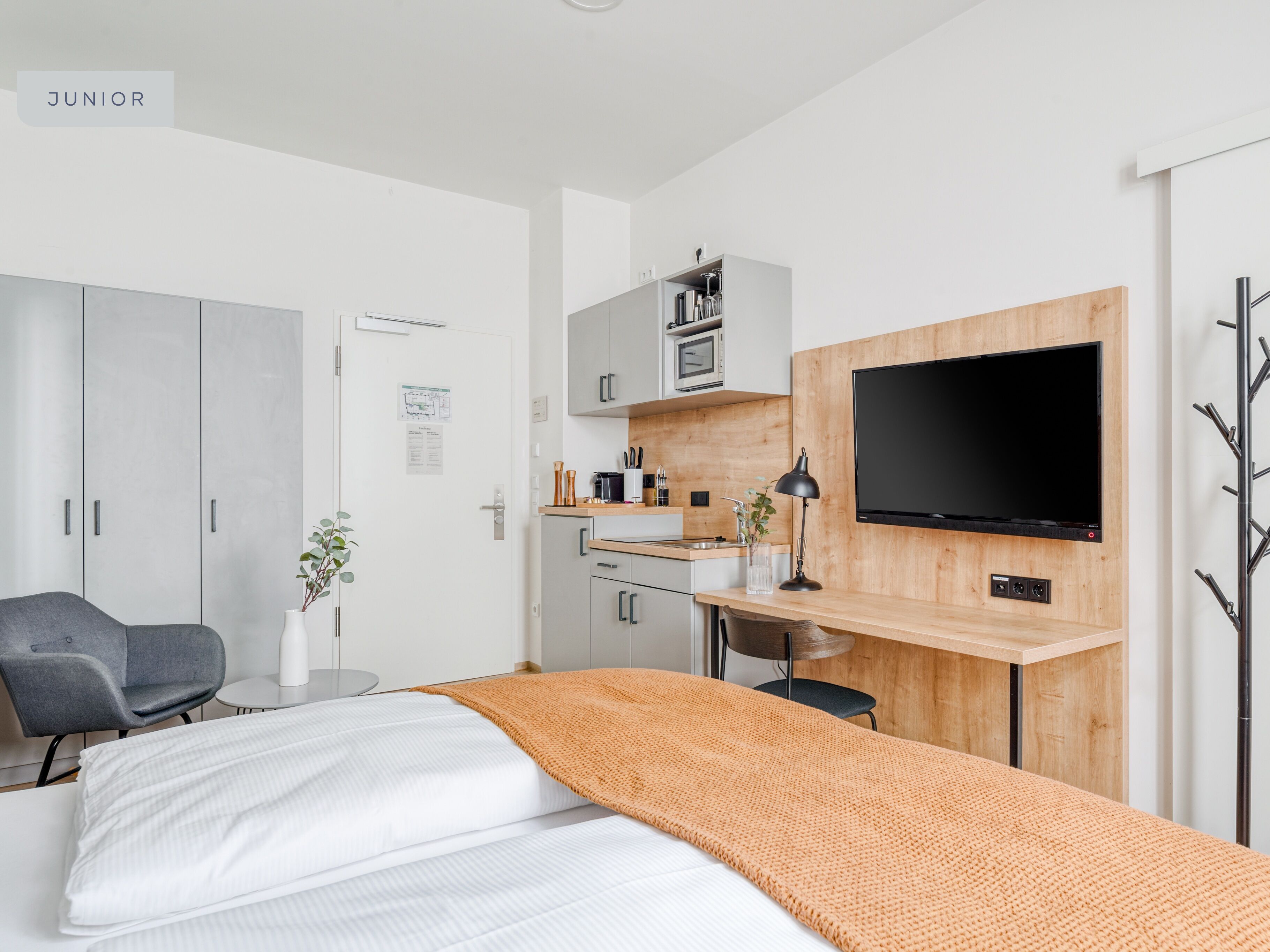 A modern junior room with a bed, a desk, a TV, and a kitchenette.