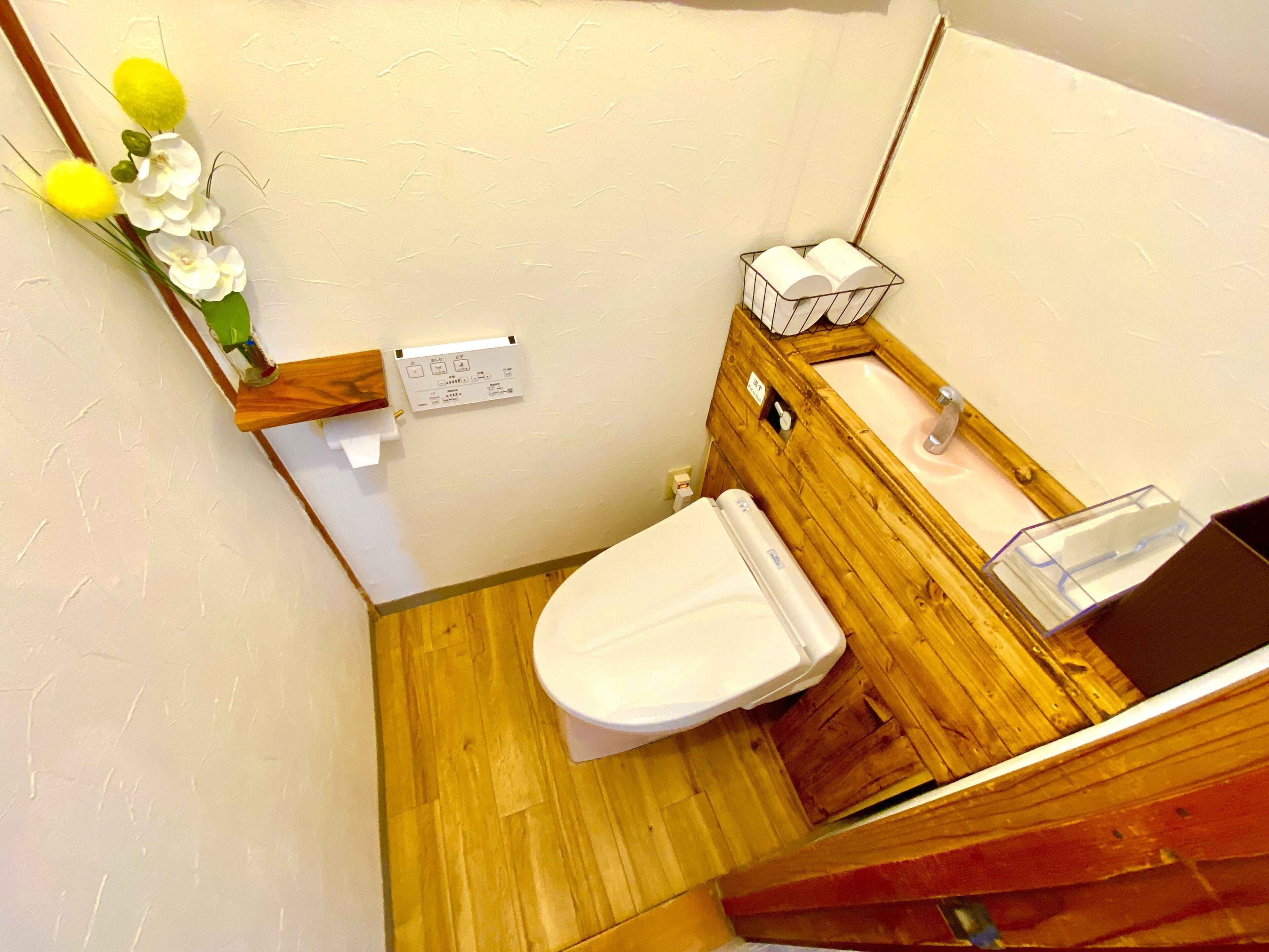A small bathroom with a wooden floor, a white toilet, a wooden vanity with a mirror, and a small shelf with toiletries.