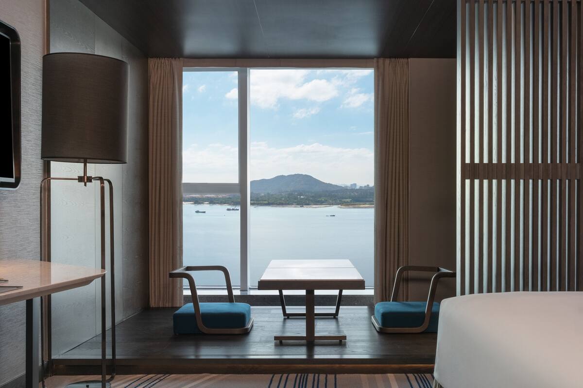 A hotel room with a large window offering a view of a body of water and a distant mountain. There is a bed, a table, and two chairs.
