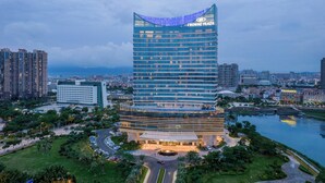 A modern hotel with a curved glass facade, named Crowne Plaza, surrounded by greenery and a body of water. - Crowne Plaza Xiamen Jimei by IHG (Xiamen)
