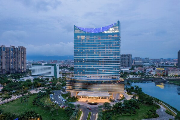 A modern hotel with a curved glass facade, named Crowne Plaza, surrounded by greenery and a body of water. - Crowne Plaza Xiamen Jimei by IHG (Xiamen)