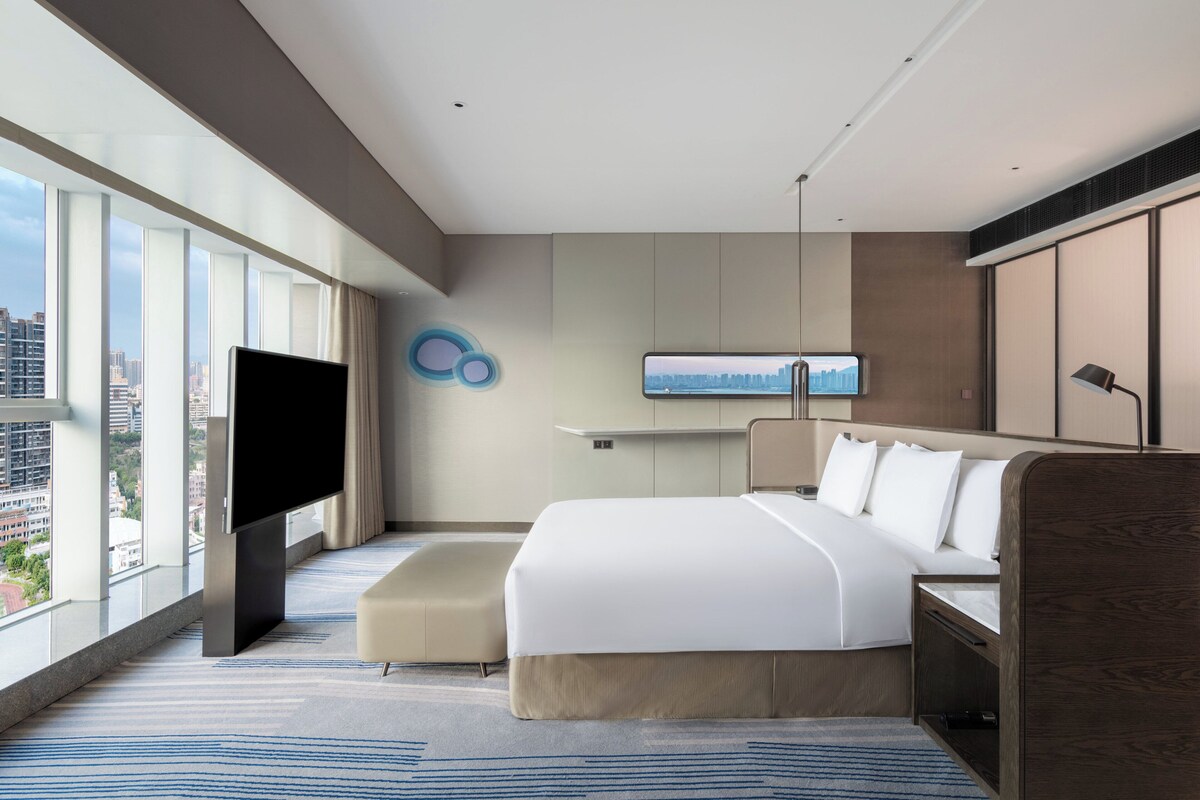 A modern hotel room with a large bed, a flat-screen TV, a city view, and a minimalist design.