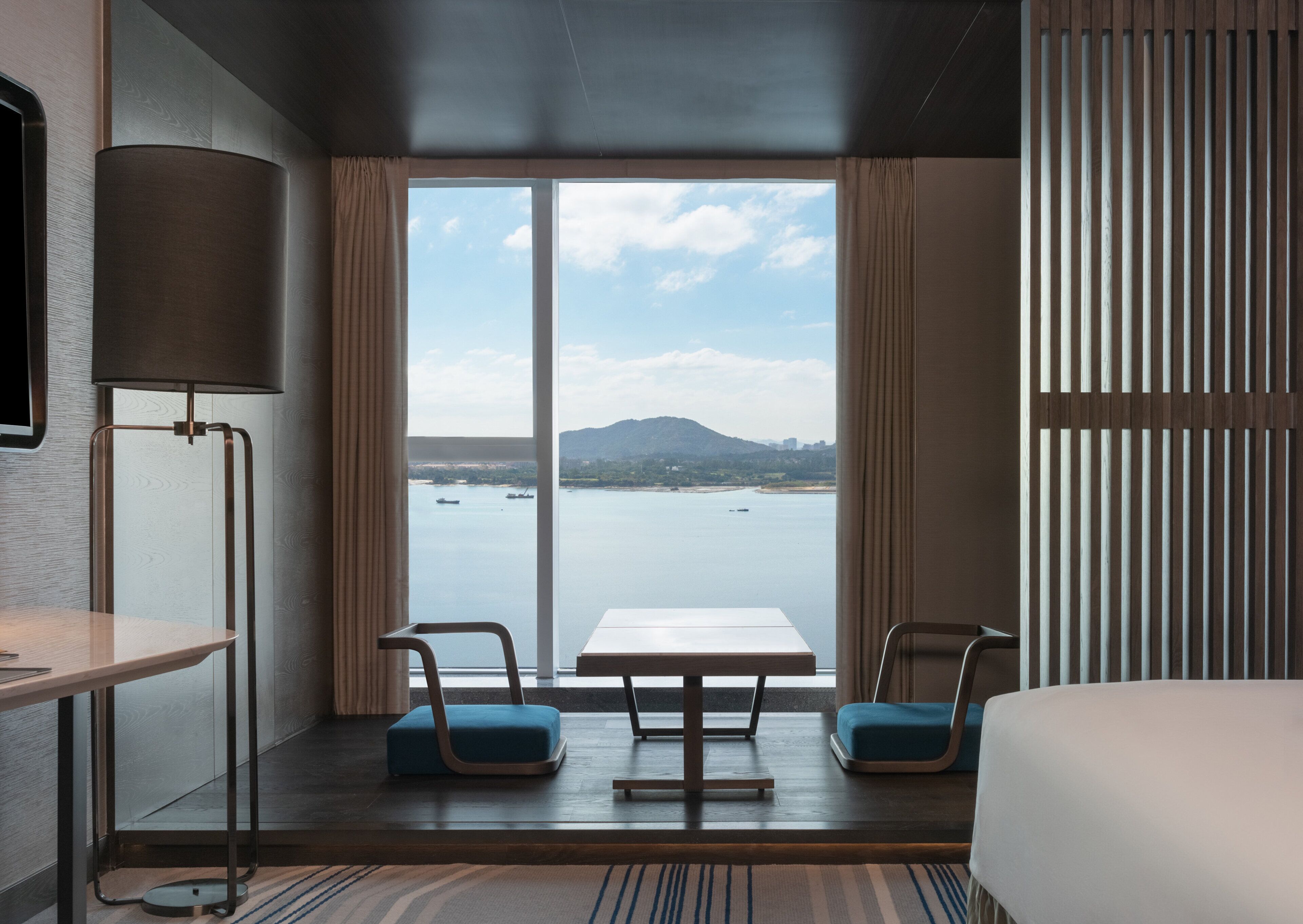 A hotel room with a large window, a bed, a table, two chairs, and a lamp.