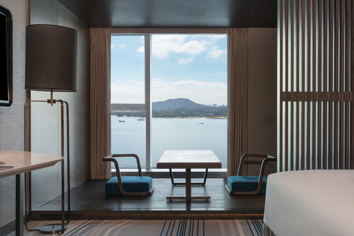 A hotel room with a large window offering a view of a body of water and a distant mountain. There is a bed, a table, and two chairs.