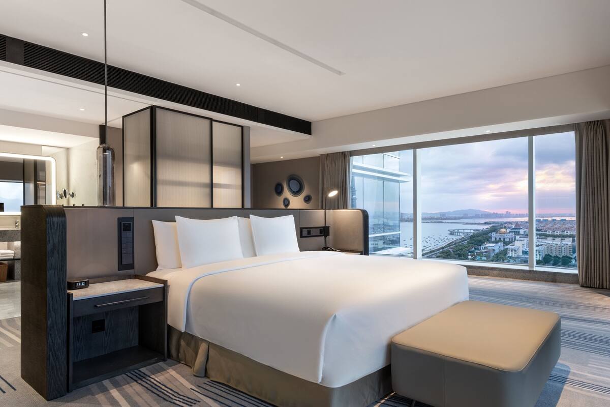 A modern hotel room with a large bed, a desk, and a view of the cityscape.