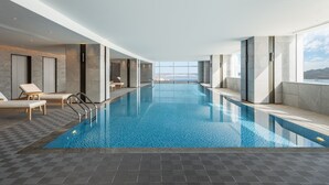 A modern indoor swimming pool with clear blue water, lounge chairs, and a view of a distant cityscape through large windows. - Crowne Plaza Xiamen Jimei by IHG (Xiamen)