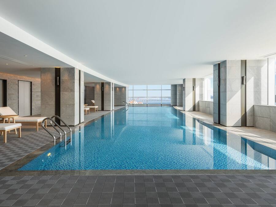 A modern indoor swimming pool with clear blue water, lounge chairs, and a view of a distant cityscape through large windows.