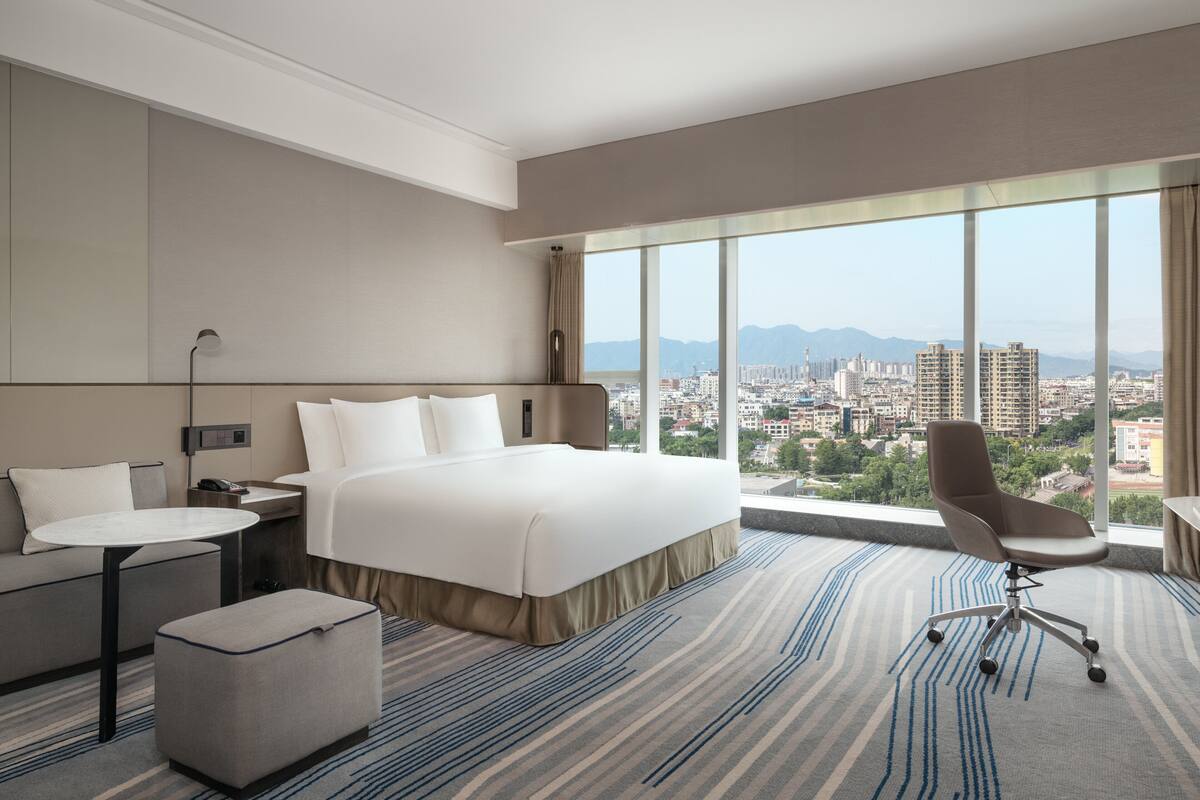 A hotel room with a large bed, a desk, a chair, and a view of the city.