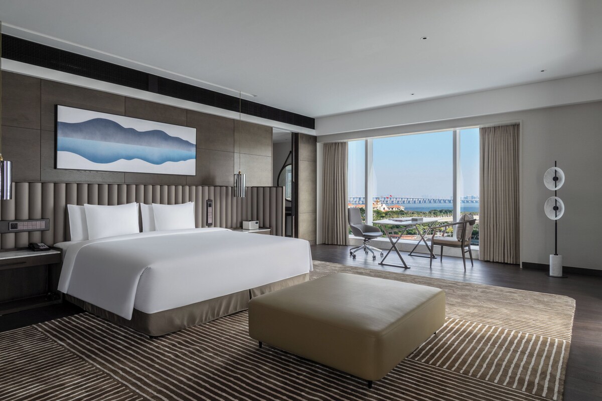 A modern hotel room with a large bed, a view of the city, and a balcony with a chair.