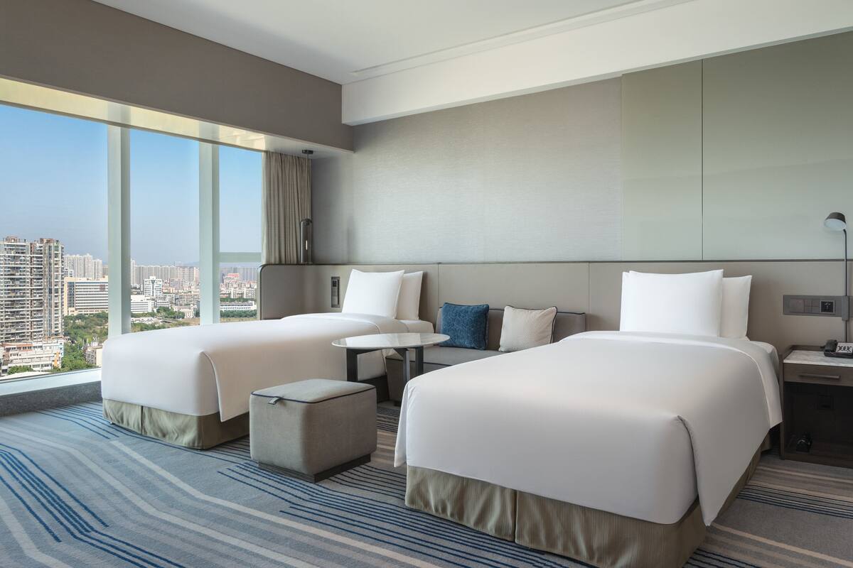 A hotel room with two beds, a small table, and a view of the city.