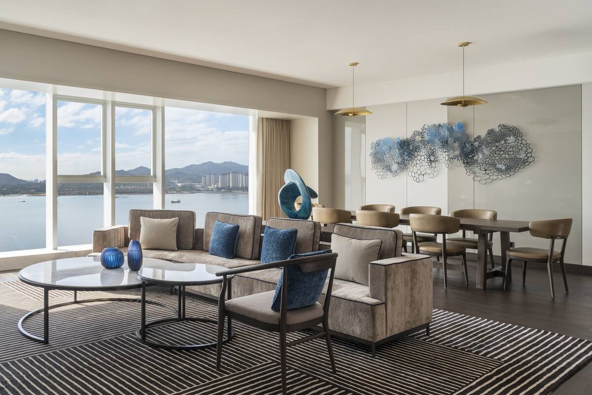 A modern hotel room with a large window offering a city view, a round dining table with chairs, and a sofa with blue cushions.